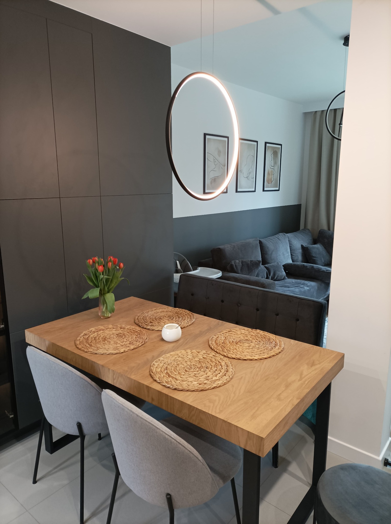 Nowoczesna jadalnia z drewnianym stołem, szarymi krzesłami i okrągłym, wiszącym oświetleniem. Na stole wazon z tulipanami i plecione podkładki. W tle szara zabudowa kuchenna i sofa.