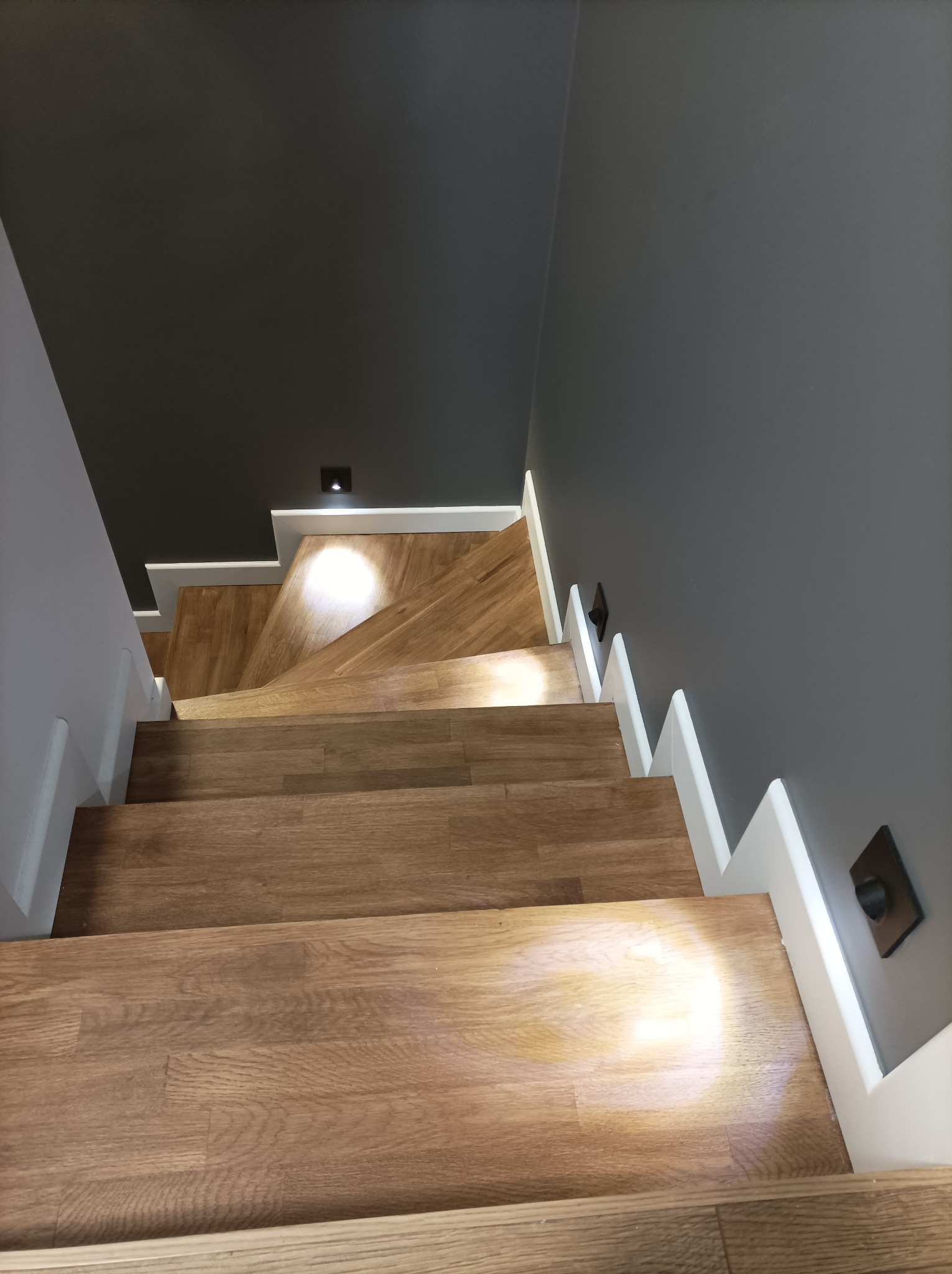 Drewniane schody z białymi balustradami i szarą ścianą, oświetlone punktowym światłem z góry, widok z góry.