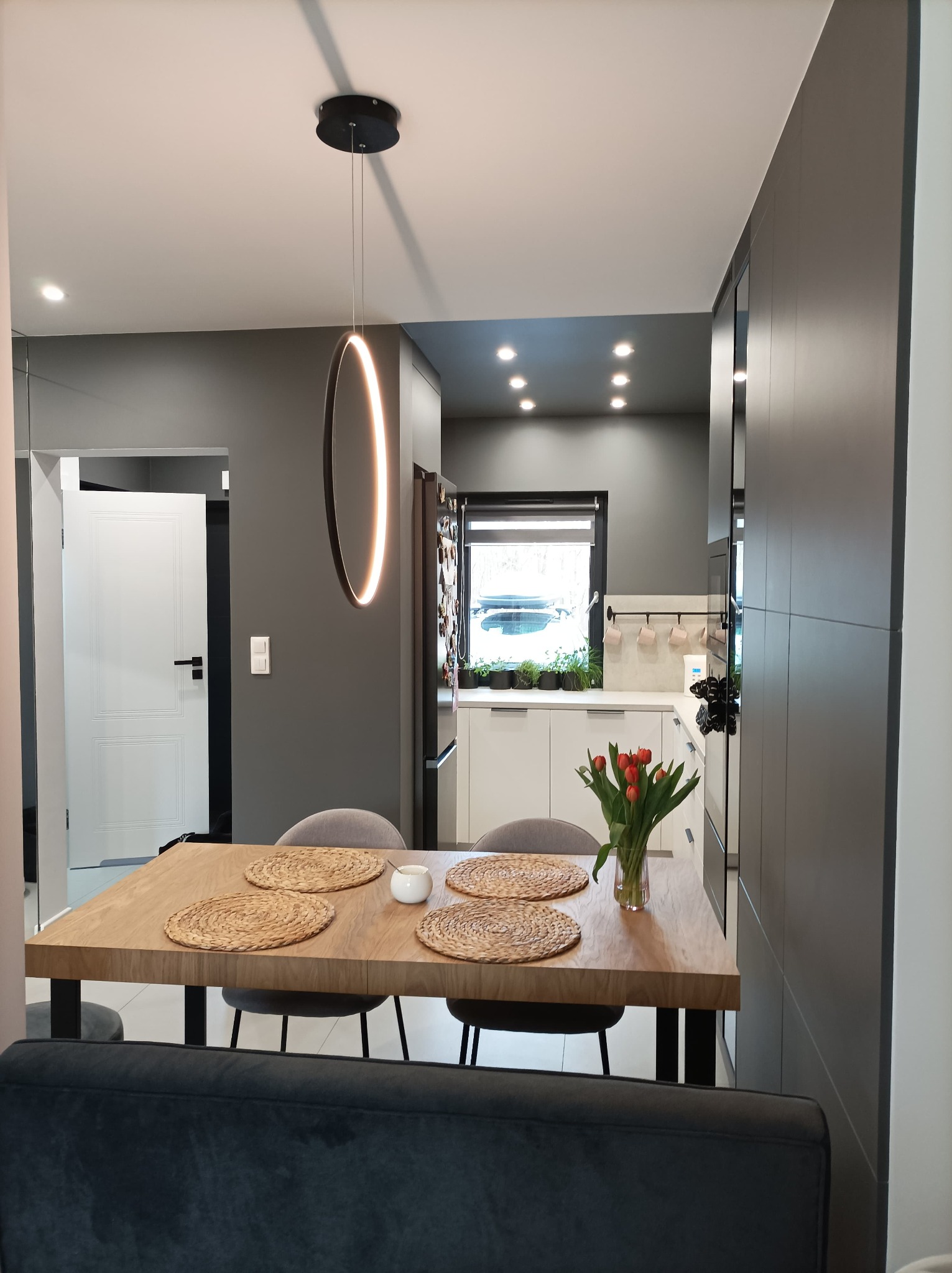 Nowoczesna kuchnia z ciemnoszarymi ścianami, drewnianym stołem z okrągłymi podkładkami i bukietem czerwonych tulipanów, oświetlona okrągłą lampą LED i punktowymi światłami w suficie.