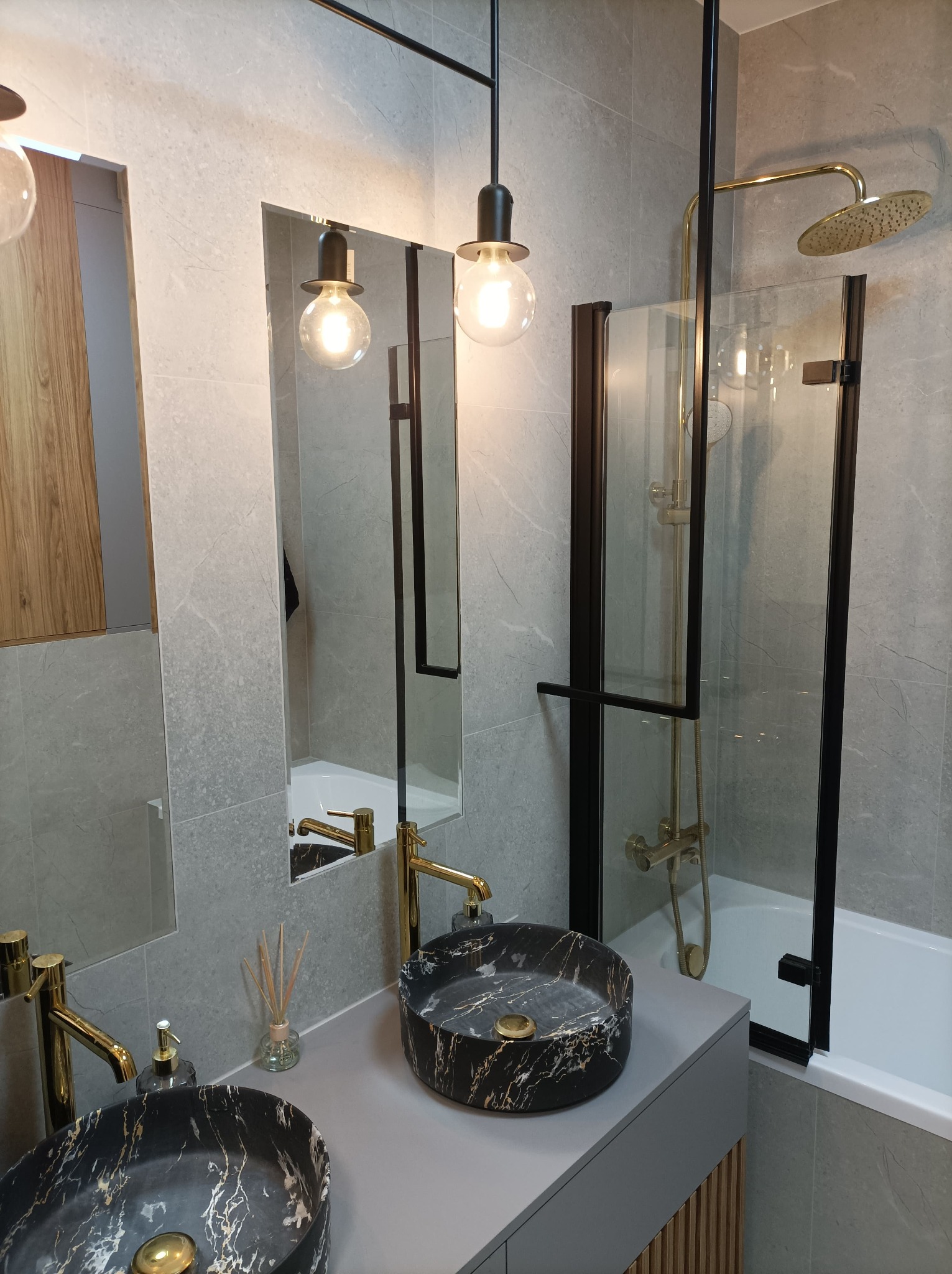 Nowoczesna łazienka z dwoma marmurkowymi umywalkami, złotymi bateriami, szklaną kabiną prysznicową z czarną ramą oraz wiszącymi lampami z żarówkami Edisona.
