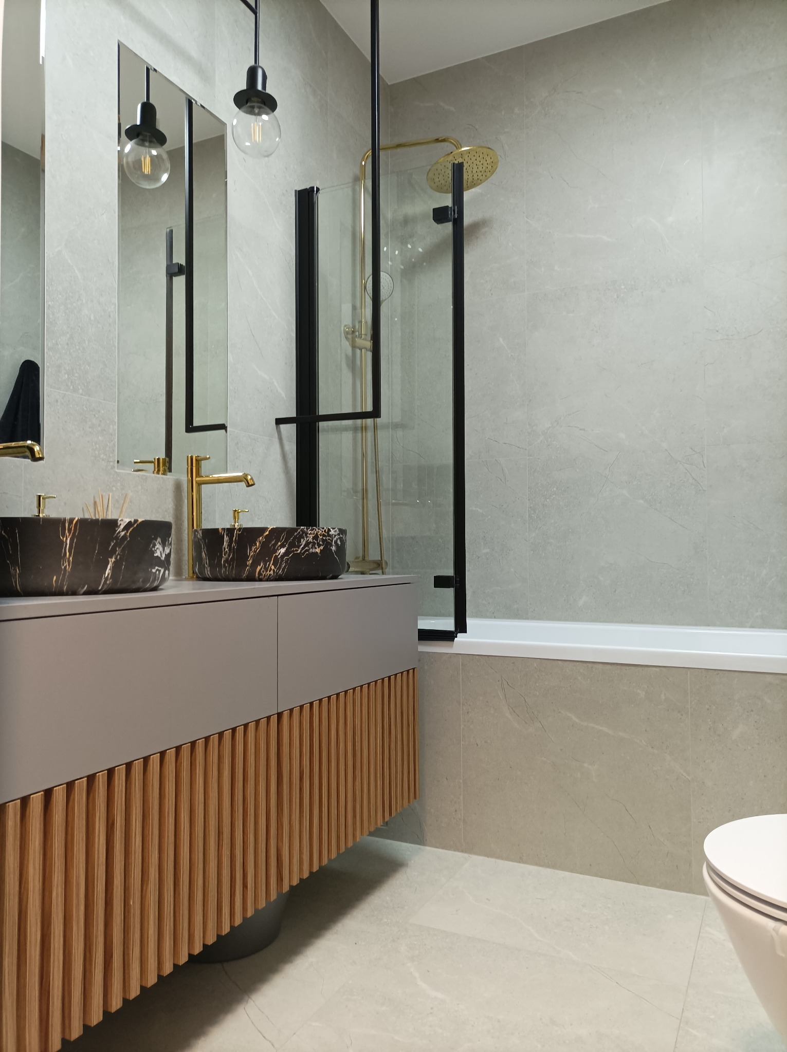 Nowoczesna łazienka z marmurowymi umywalkami nablatowymi, złotą armaturą, kabiną prysznicową z czarnymi profilami i drewnianą szafką pod umywalką z lamelami.