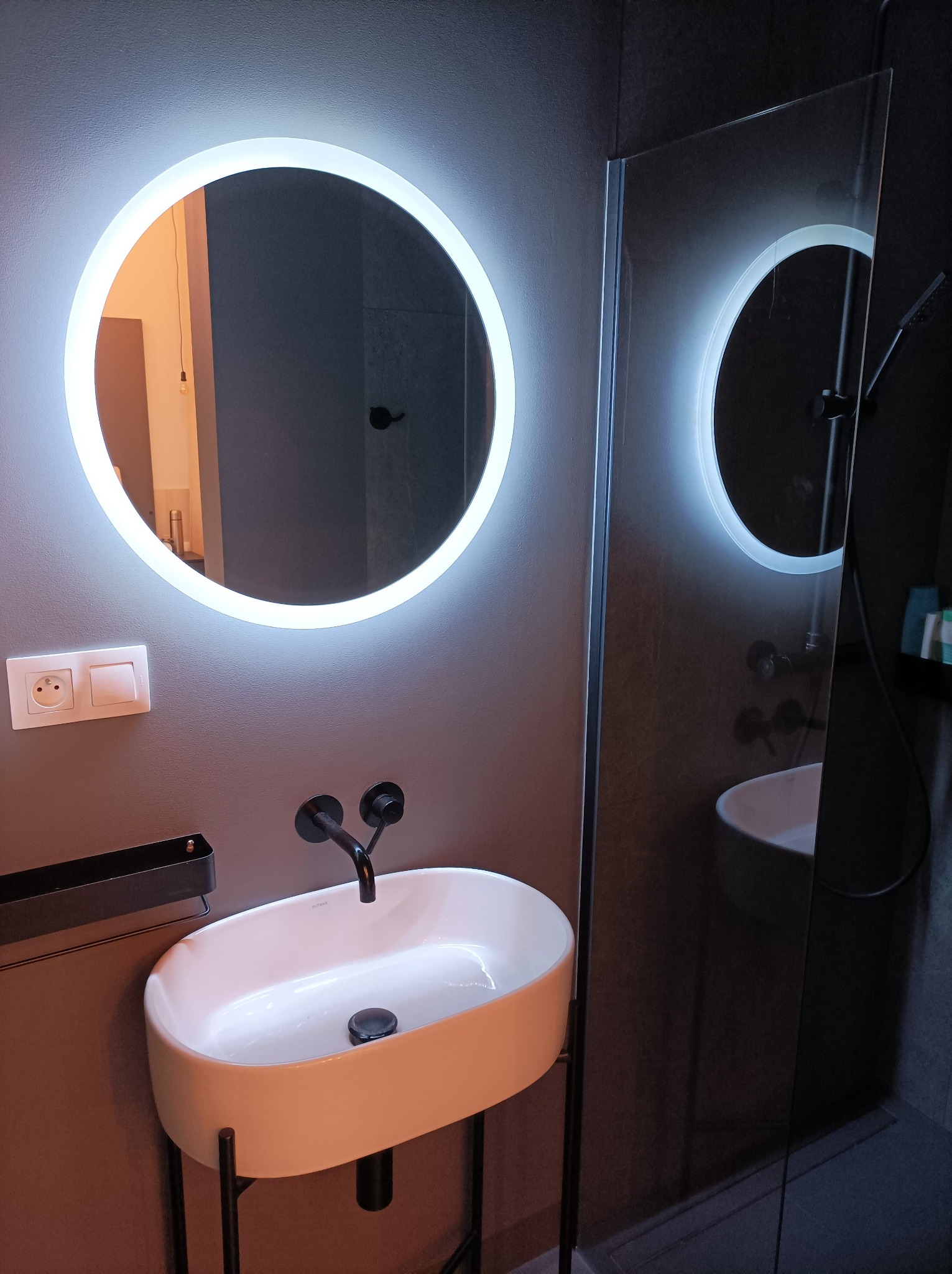 Nowoczesna łazienka z owalną umywalką na czarnym stelażu, okrągłym lustrem z podświetleniem LED i kabiną prysznicową ze szklaną ścianką.