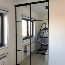 Przesuwne drzwi w stylu loft z czarnymi szprosami i lustrzanymi panelami, odbijające wnętrze pokoju z wiszącym fotelem i grzejnikiem.