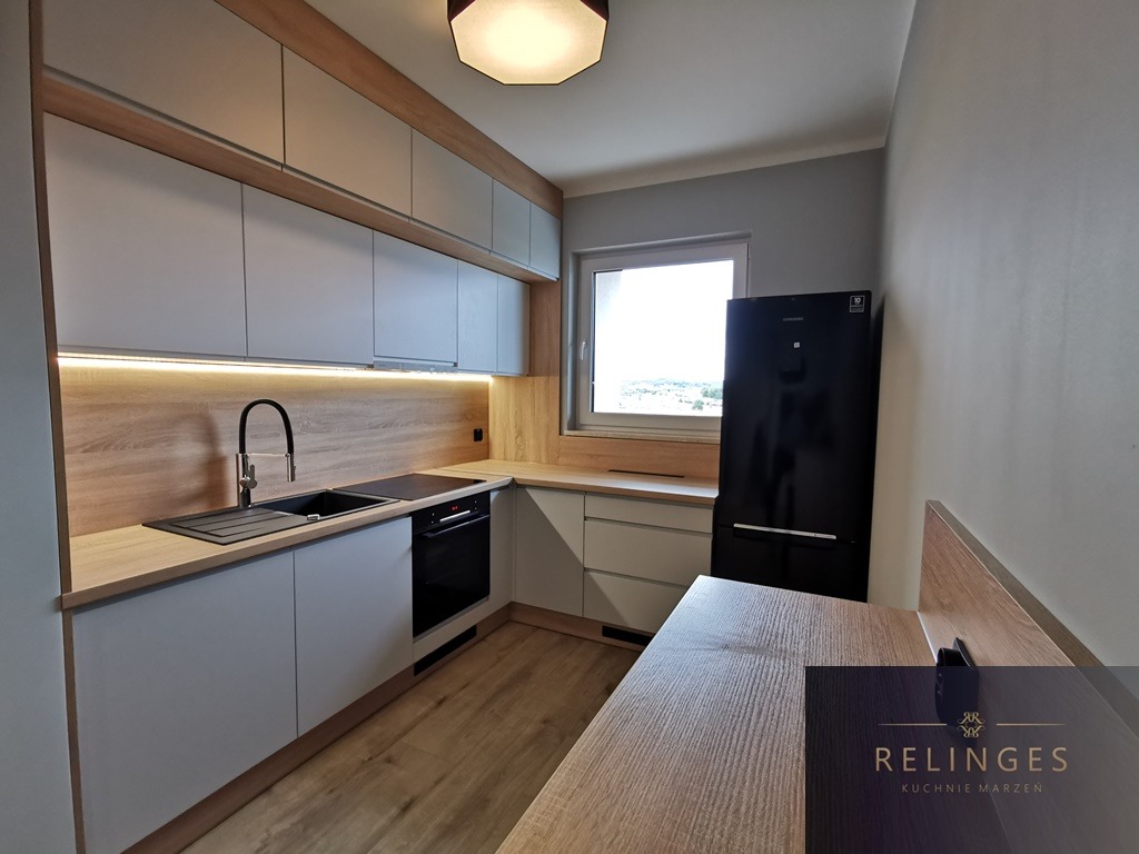 Jasnoszara kuchnia z drewnianymi akcentami, czarnym zlewem i baterią, oraz czarną lodówką obok okna, oświetlona listwą LED podszafkową.