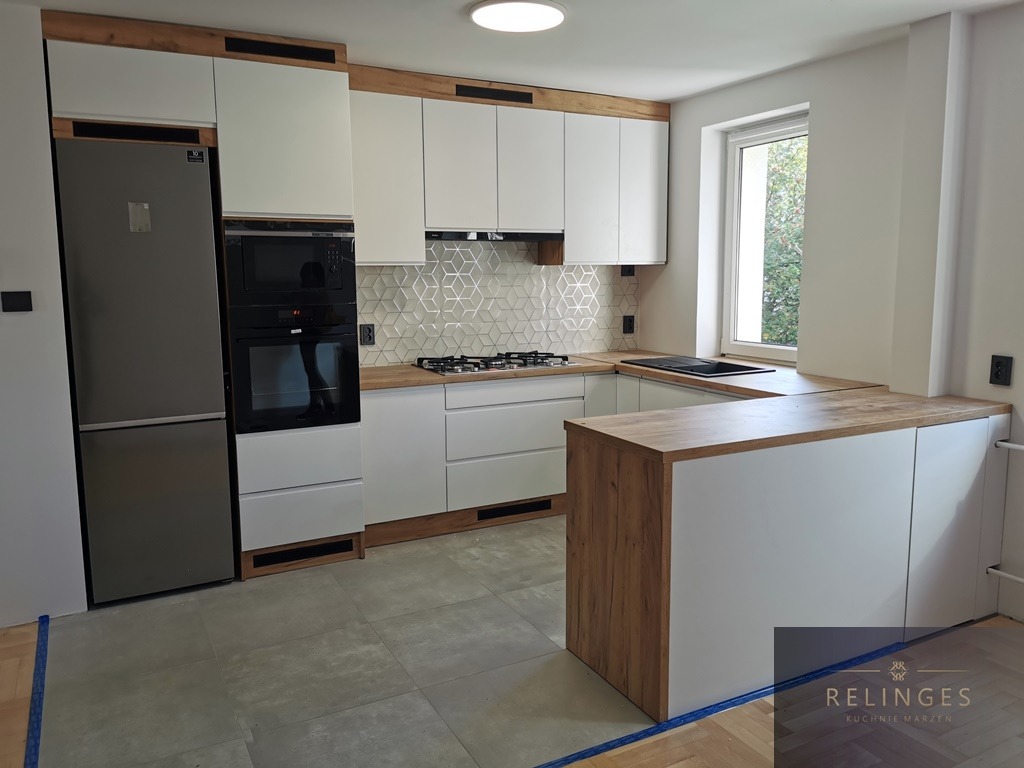 Nowoczesna, jasna kuchnia z białymi frontami i drewnianymi akcentami, wykończona geometryczną mozaiką nad blatem, z widocznym oknem i wyspą kuchenną.