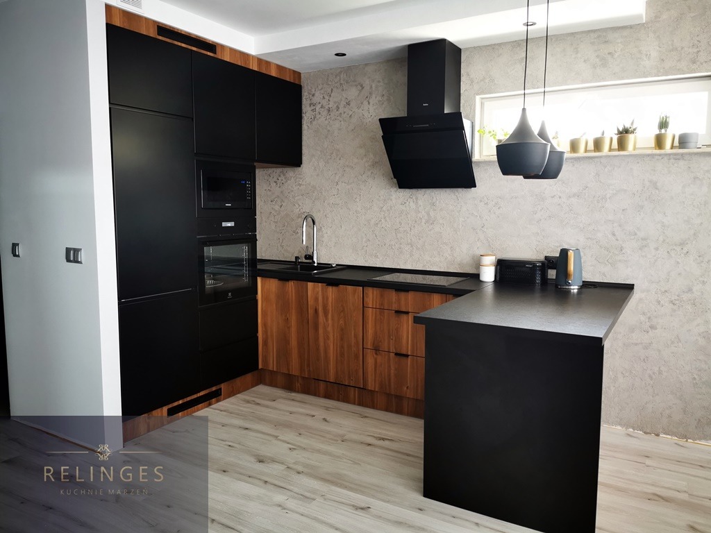 Nowoczesna kuchnia z czarnymi frontami szafek i drewnianymi elementami, wyspa kuchenna z czarnym blatem, szara ściana z dekoracyjnym tynkiem, wiszące lampy i okno z roślinami.