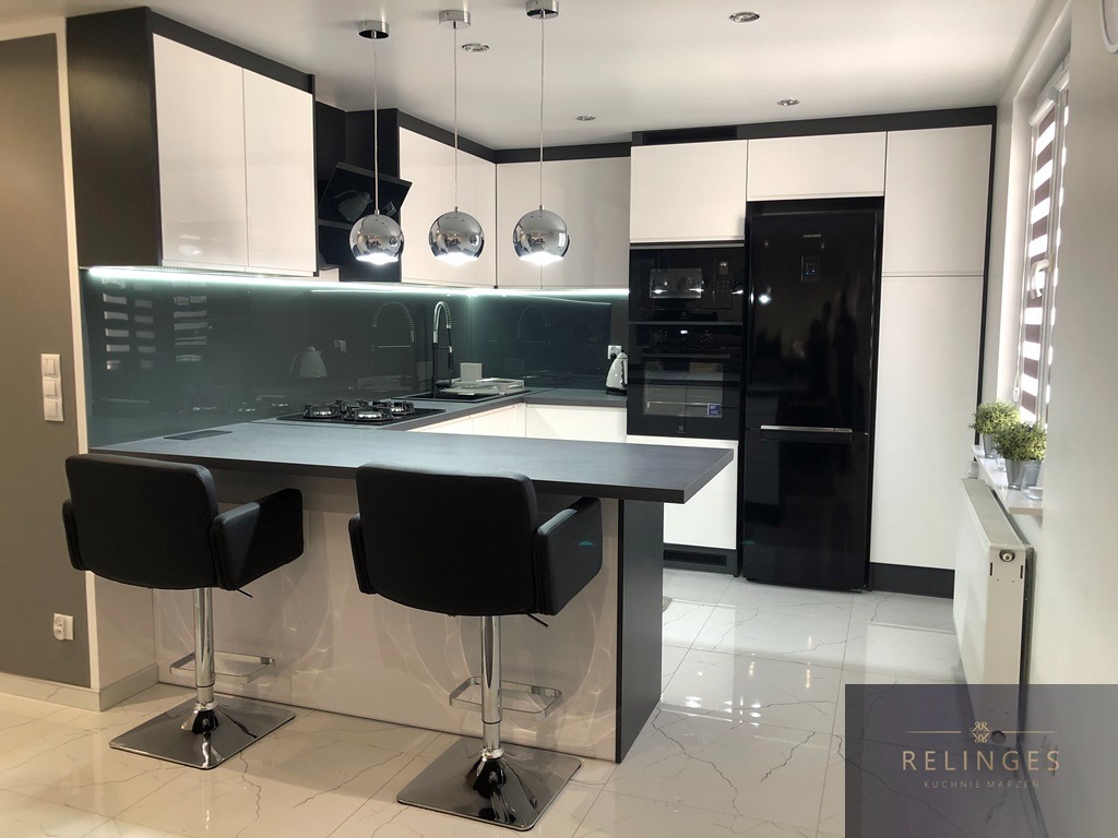 Nowoczesna kuchnia z białymi szafkami z czarnymi akcentami, szarym blatem i szklanym panelem nad blatem, oświetlona trzema kulistymi lampami wiszącymi nad wyspą z dwoma czarnymi hokerami.