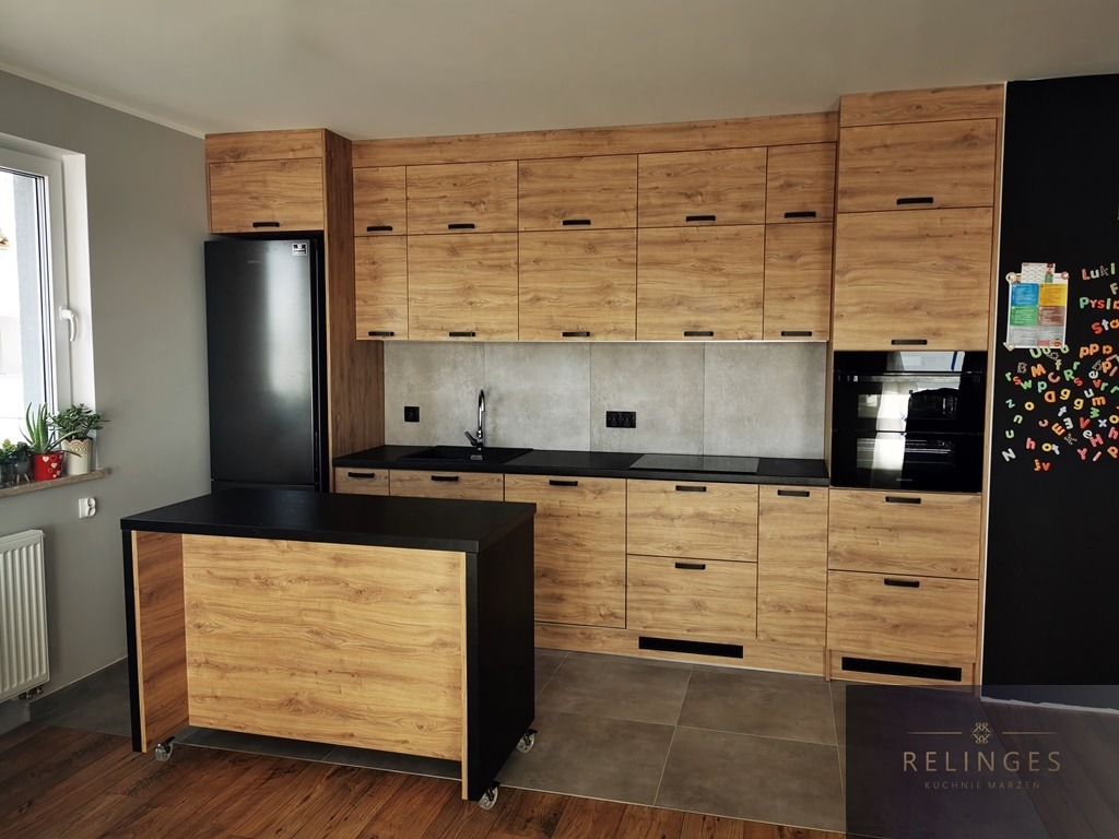 Kompletna zabudowa kuchenna z frontami imitującymi drewno, czarnym blatem i wyspą na kółkach, widoczna czarna lodówka i tablica kredowa z magnesami.