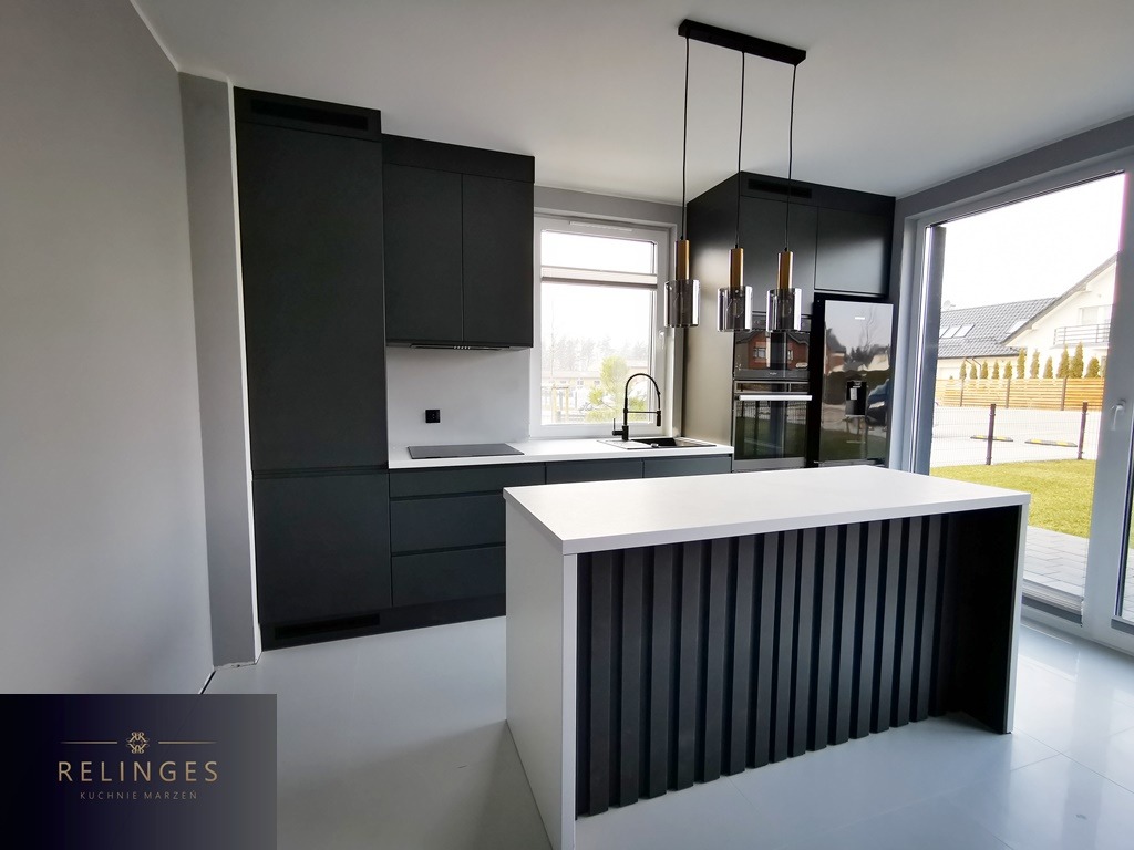 Nowoczesna kuchnia w odcieniach szarości z wyspą o ryflowanym froncie i wiszącymi lampami, widok na ogród przez duże okno.