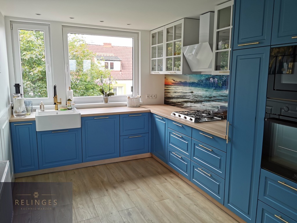 Niebieska kuchnia z frezowanymi frontami, drewnianym blatem i białym zlewem farmerskim pod oknem z widokiem na zieleń. Nad blatem panel szklany z motywem morskim.