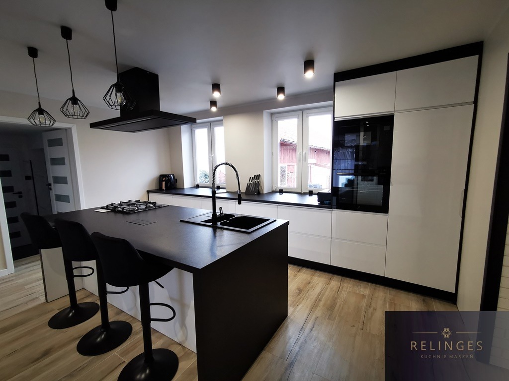 Nowoczesna kuchnia z czarnym blatem wyspowym, białymi szafkami i drewnianą podłogą, oświetlona trzema wiszącymi lampami w stylu industrialnym, z widocznym logo firmy w prawym dolnym rogu.