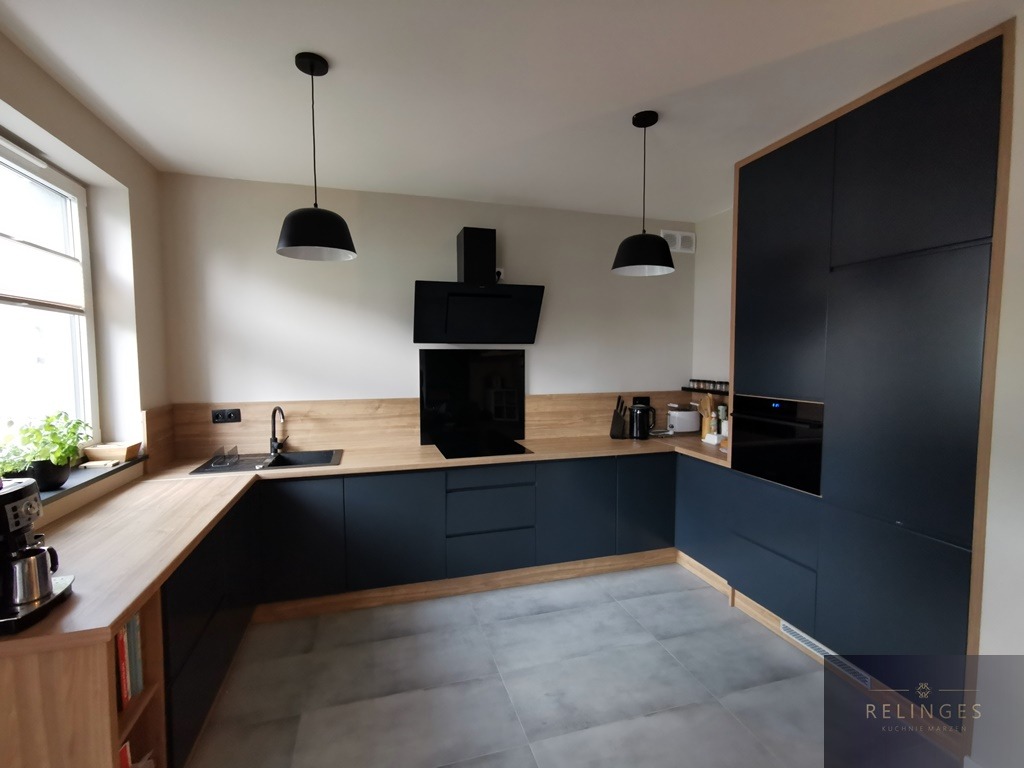 Nowoczesna kuchnia w odcieniach granatu i drewna, z czarnymi lampami wiszącymi i szarą podłogą, akcentem jest drewniana półka na książki w zabudowie kuchennej.