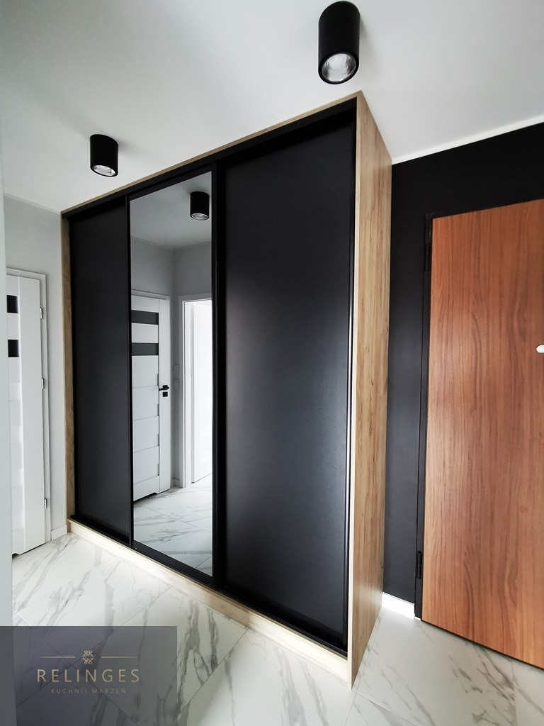 Nowoczesna szafa wnękowa z przesuwnymi drzwiami, czarne matowe fronty, obudowa w odcieniu jasnego drewna, widoczne elementy oświetlenia sufitowego oraz fragment innego pomieszczenia w odbiciu lustra.
