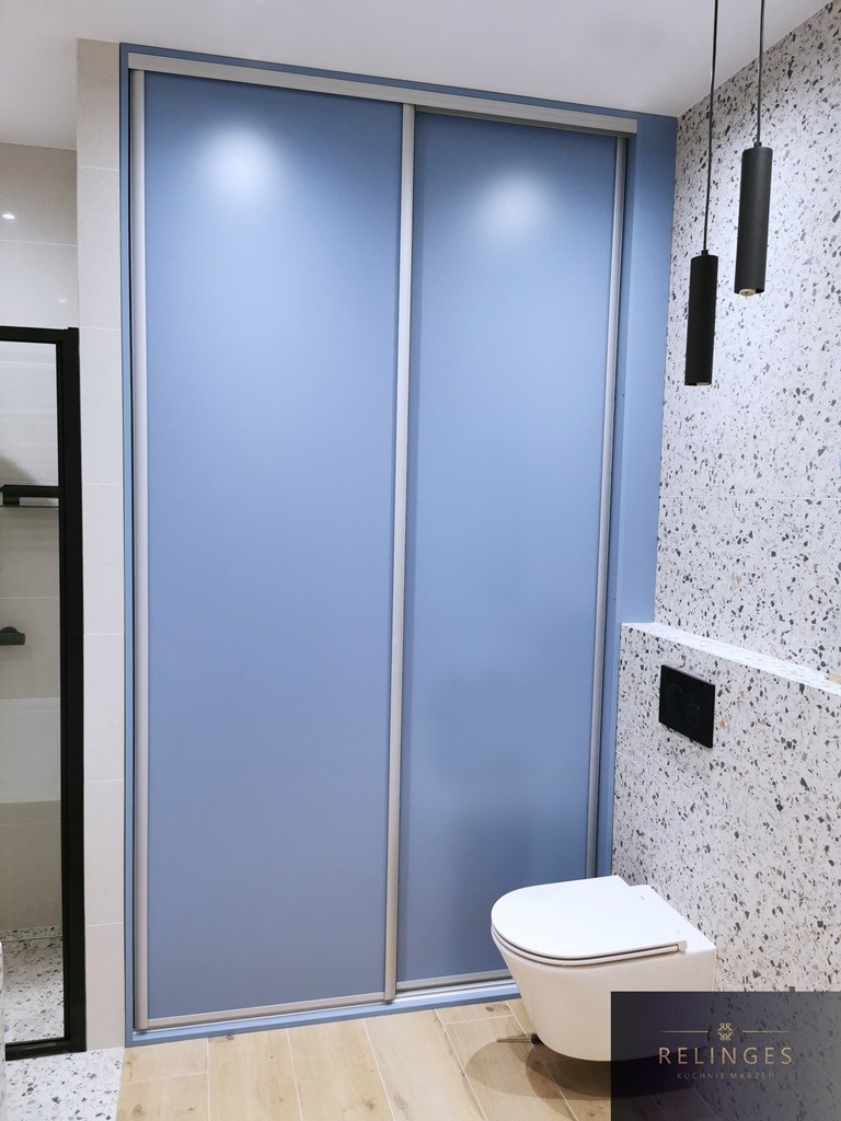 Niebieska szafa przesuwna z lustrzanymi drzwiami w nowoczesnej łazience w Elblągu, z wiszącą toaletą i lampami tubami na tle ściany w terrazzo.