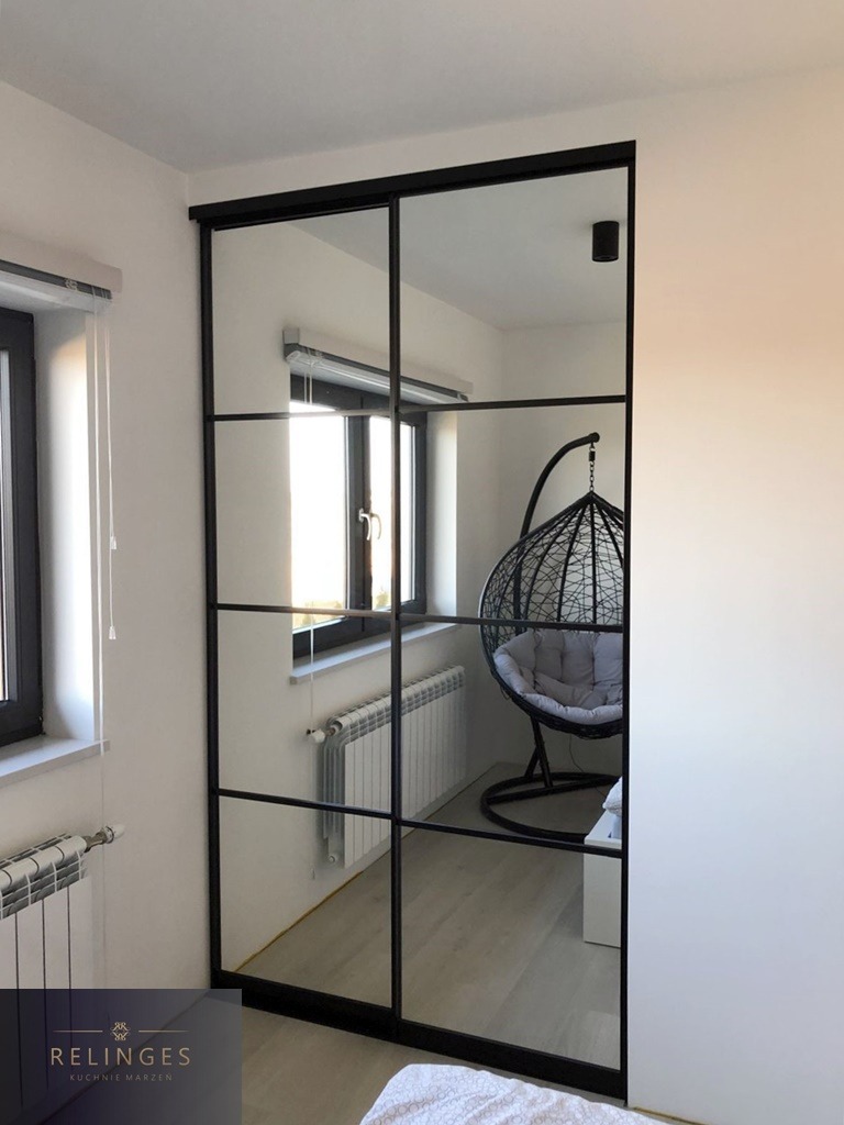 Przesuwne drzwi w stylu loft z czarnymi szprosami i lustrzanymi panelami, odbijające wnętrze pokoju z wiszącym fotelem i grzejnikiem.