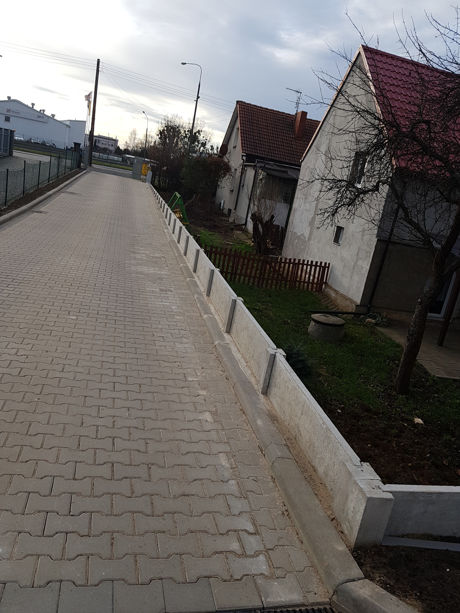 Nowo wybudowany, surowy, betonowy murek oddzielający chodnik z kostki brukowej od terenu posesji z widocznymi zabudowaniami w tle.