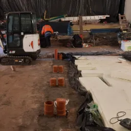 Przygotowanie podłoża pod instalację ogrzewania podłogowego: mini koparka Bobcat E19 z młotem pneumatycznym, ułożone płyty izolacyjne styropianowe Finnfoam, rury kanalizacyjne i siatka zbrojeniowa...