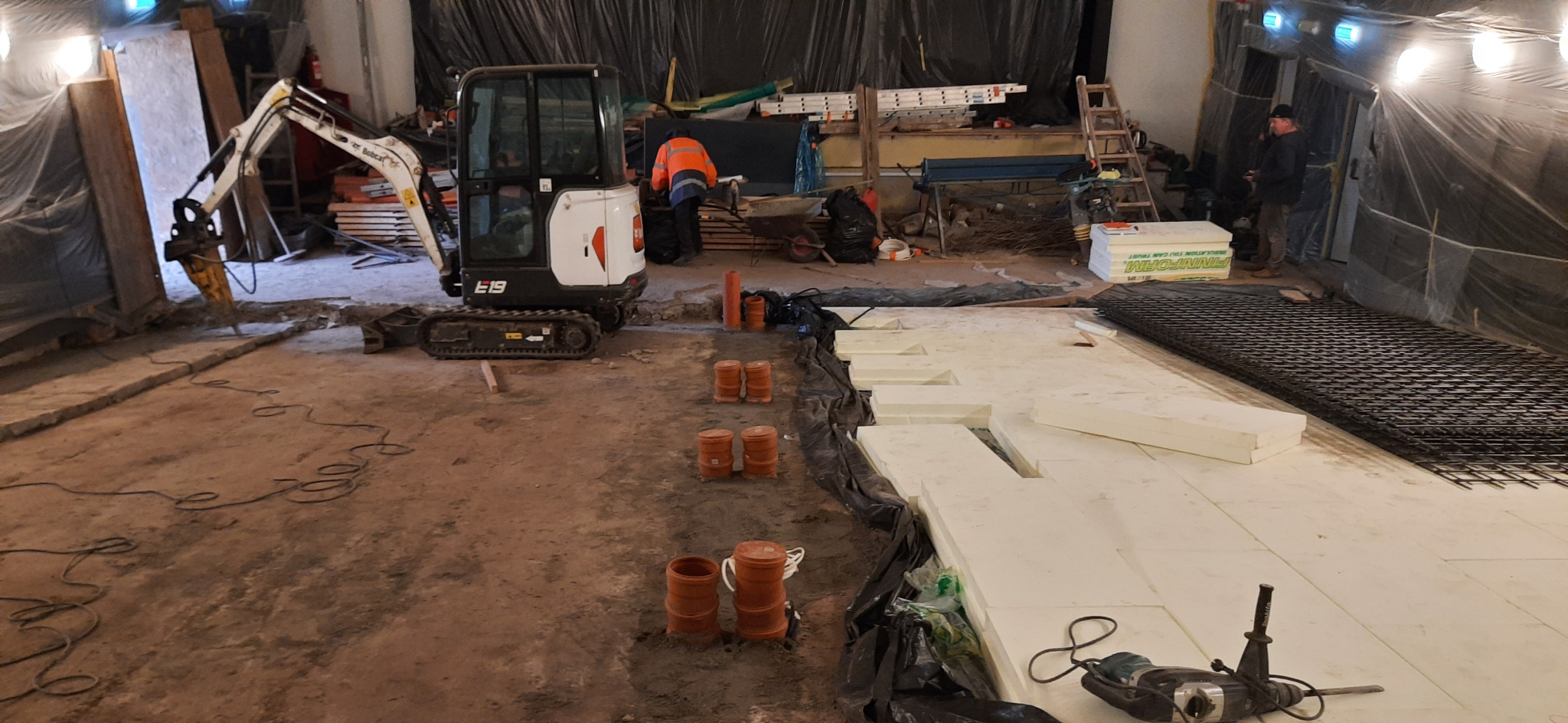 Przygotowanie podłoża pod instalację ogrzewania podłogowego: mini koparka Bobcat E19 z młotem pneumatycznym, ułożone płyty izolacyjne styropianowe Finnfoam, rury kanalizacyjne i siatka zbrojeniowa...