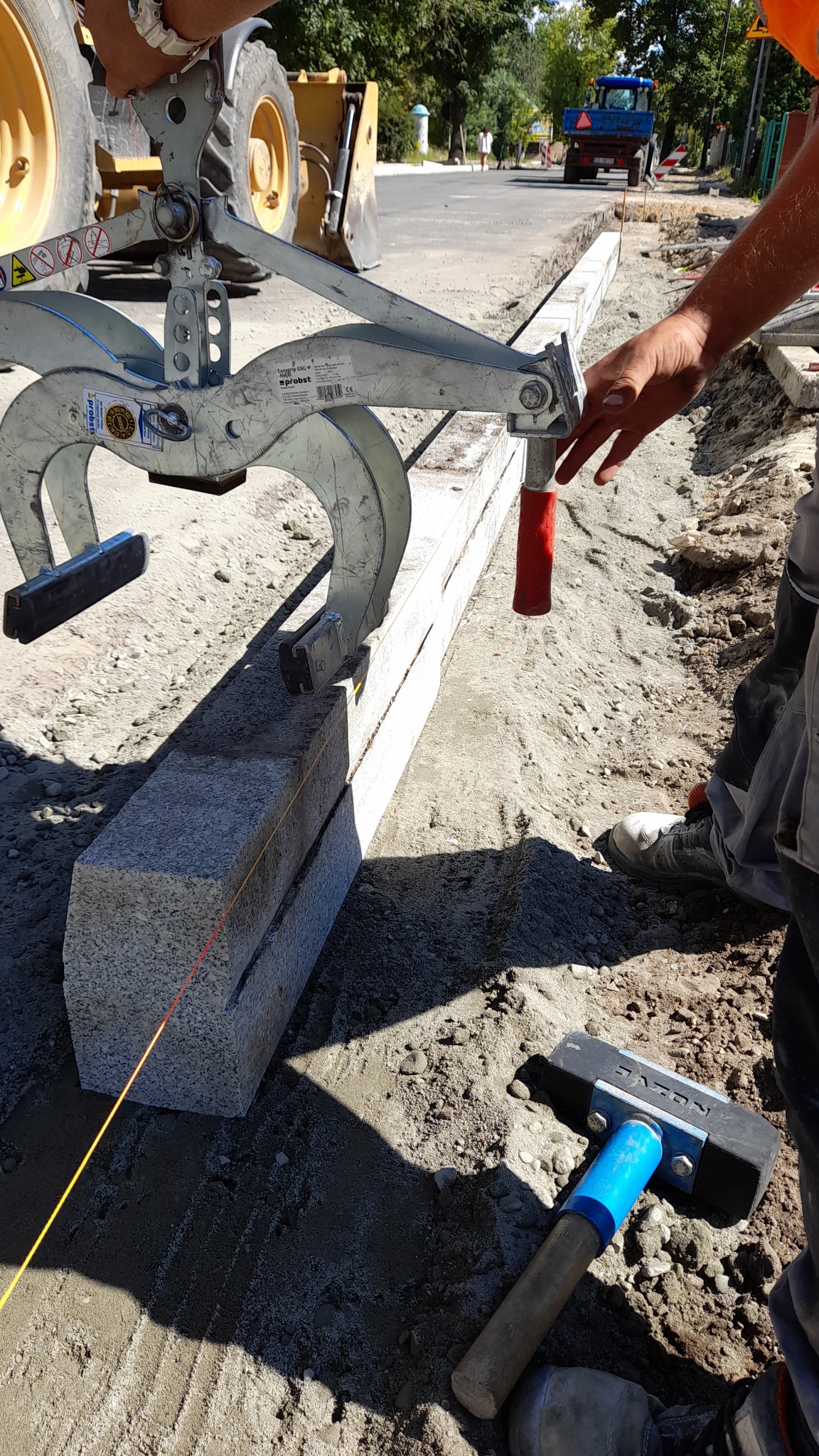 Precyzyjne układanie granitowych krawężników w Chorzowie przy użyciu chwytaka mechanicznego; widoczny sznurek traserski i gumowy młotek.