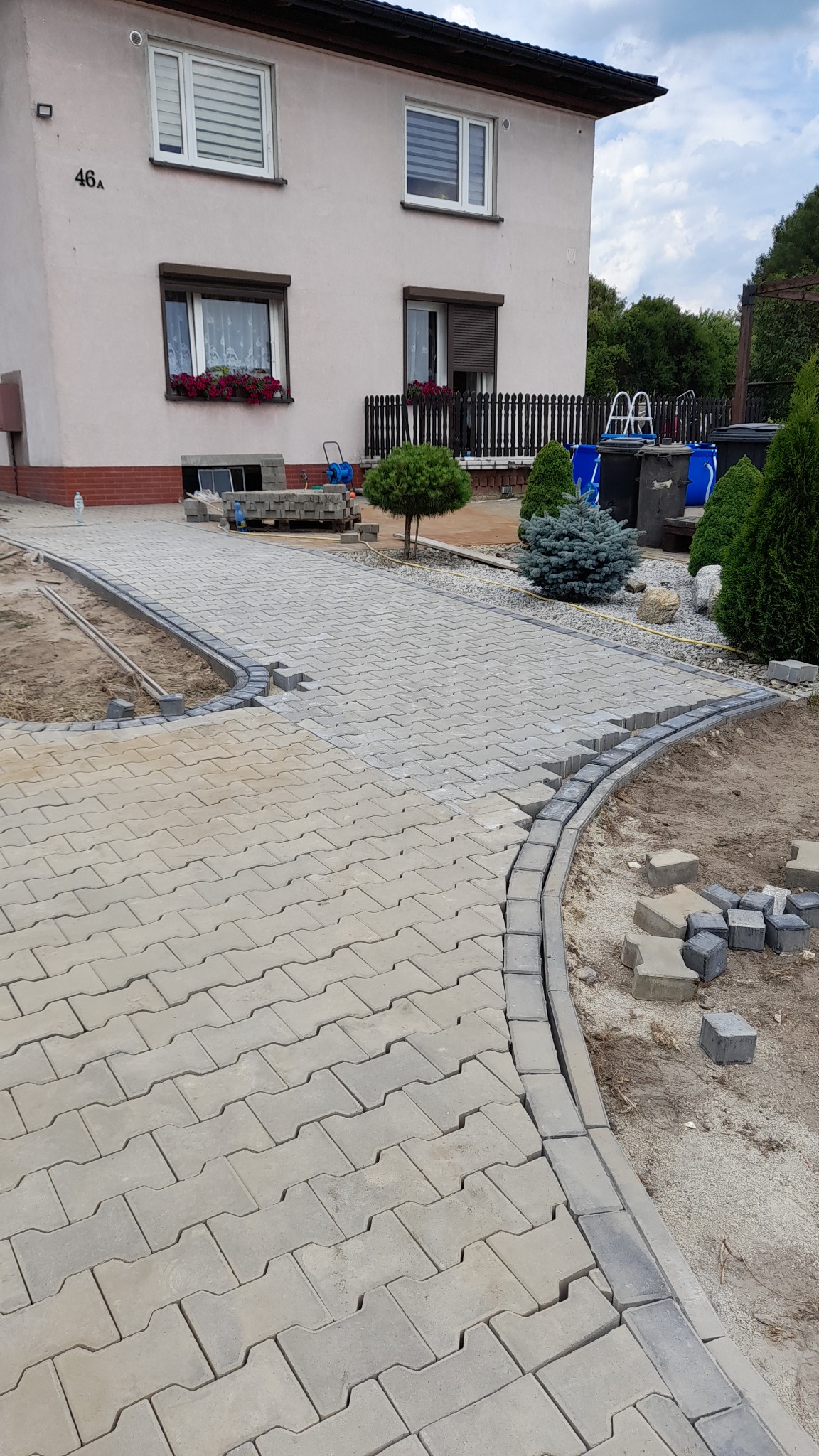 Ścieżka z jasnoszarej kostki brukowej ułożona do posesji z numerem 46A, z obrzeżem z ciemniejszej kostki, w tle widoczny trawnik, drzewa i basen.