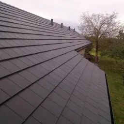 Nowoczesny, ciemnoszary dach z płaskiej dachówki ceramicznej, widok z góry, kominki wentylacyjne, drzewo w tle.