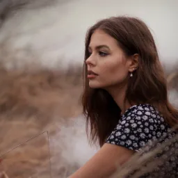 Portret młodej kobiety z profilu, w sukience w kwiaty, z kolczykami z perłami, siedzącej w trawie z dymem w tle.