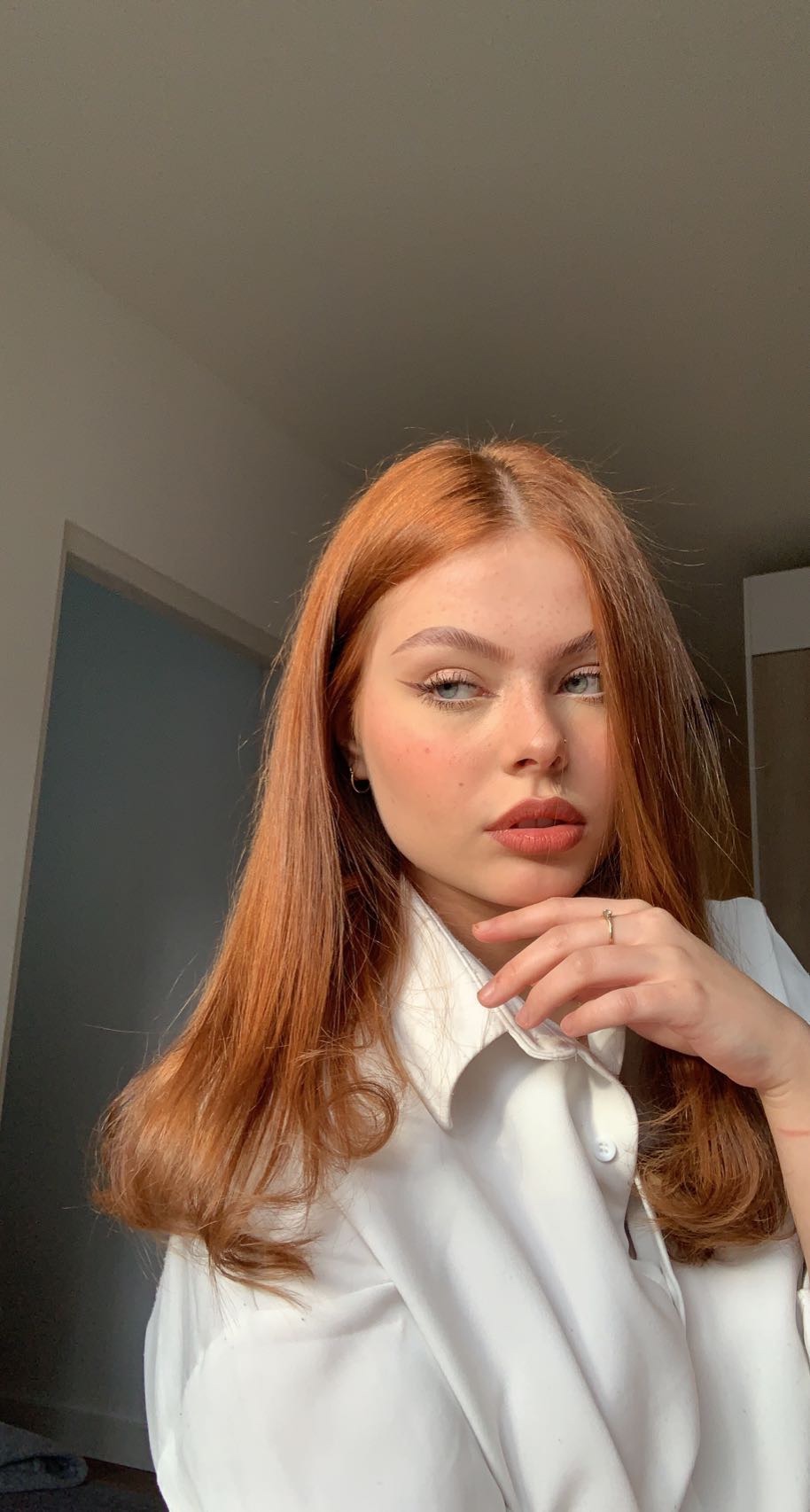Portret kobiety z rudymi włosami, w białej koszuli, z delikatnym makijażem i pierścionkiem na palcu, pozującej w naturalnym świetle.