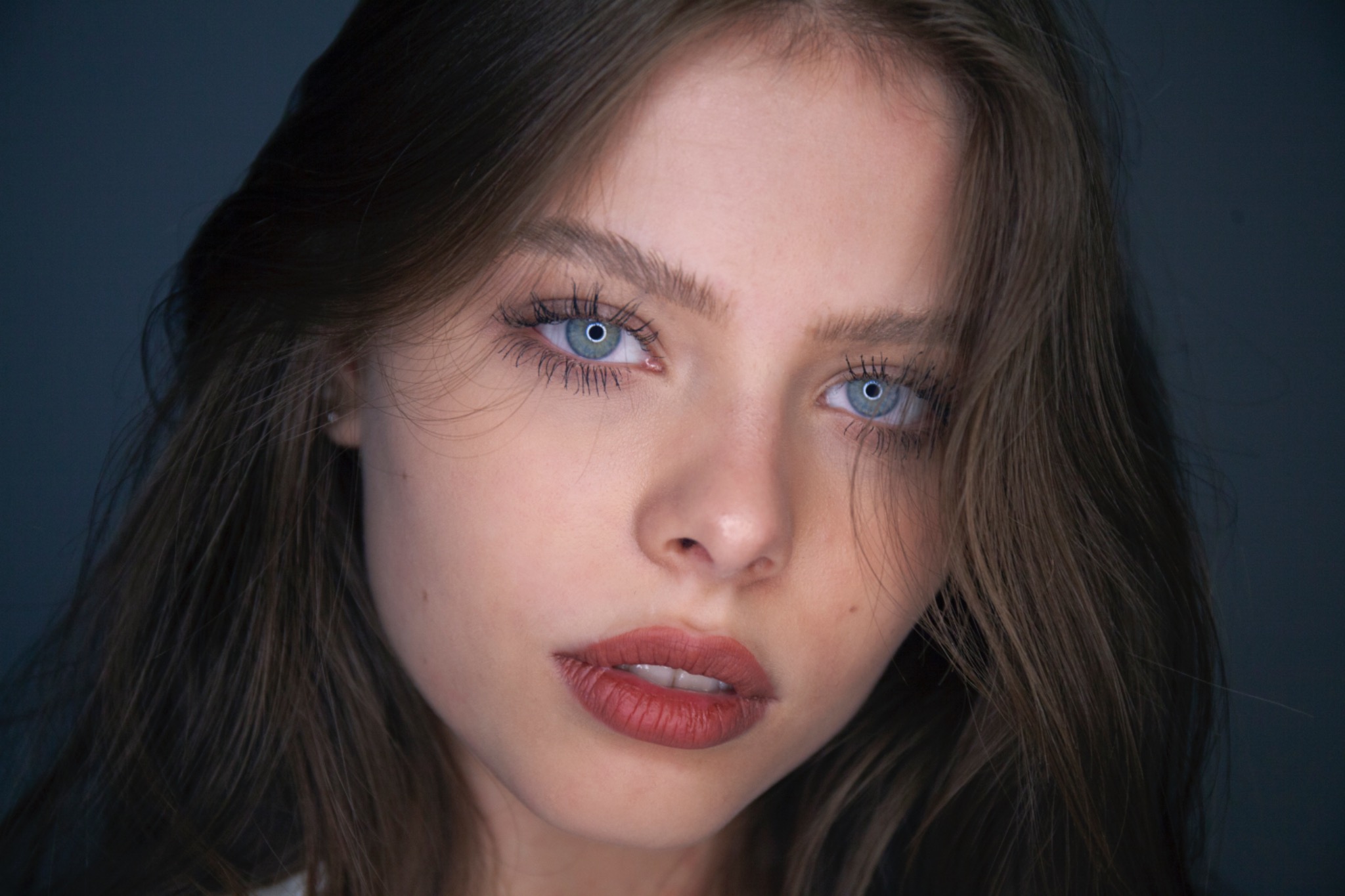 Portret młodej kobiety z jasnymi oczami, brązowymi włosami i delikatnym makijażem ust w odcieniach czerwieni na tle ciemnego tła.