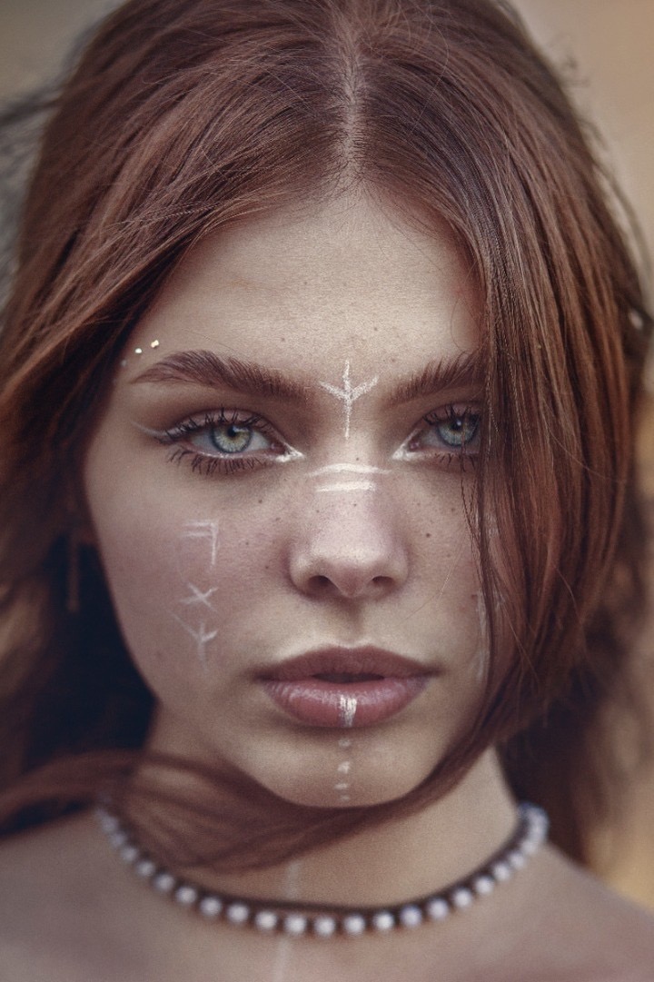 Portret młodej kobiety z rudymi włosami i jasnymi oczami, ozdobiona białymi, etnicznymi wzorami na twarzy i szyi, nosząca naszyjnik z pereł.