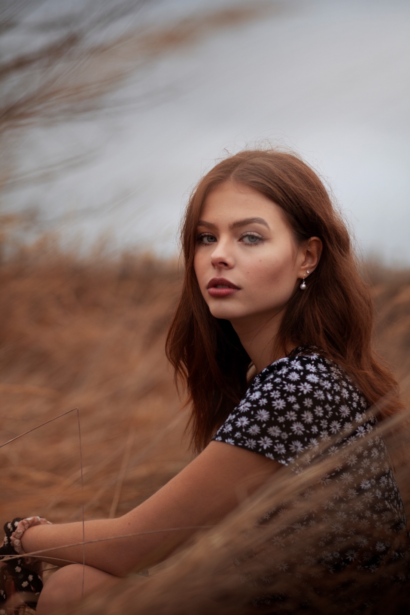 Portret młodej kobiety z rudymi włosami i kolczykiem z perłą, siedzącej w trawie, z delikatnym makijażem i wzrokiem skierowanym w stronę widza.