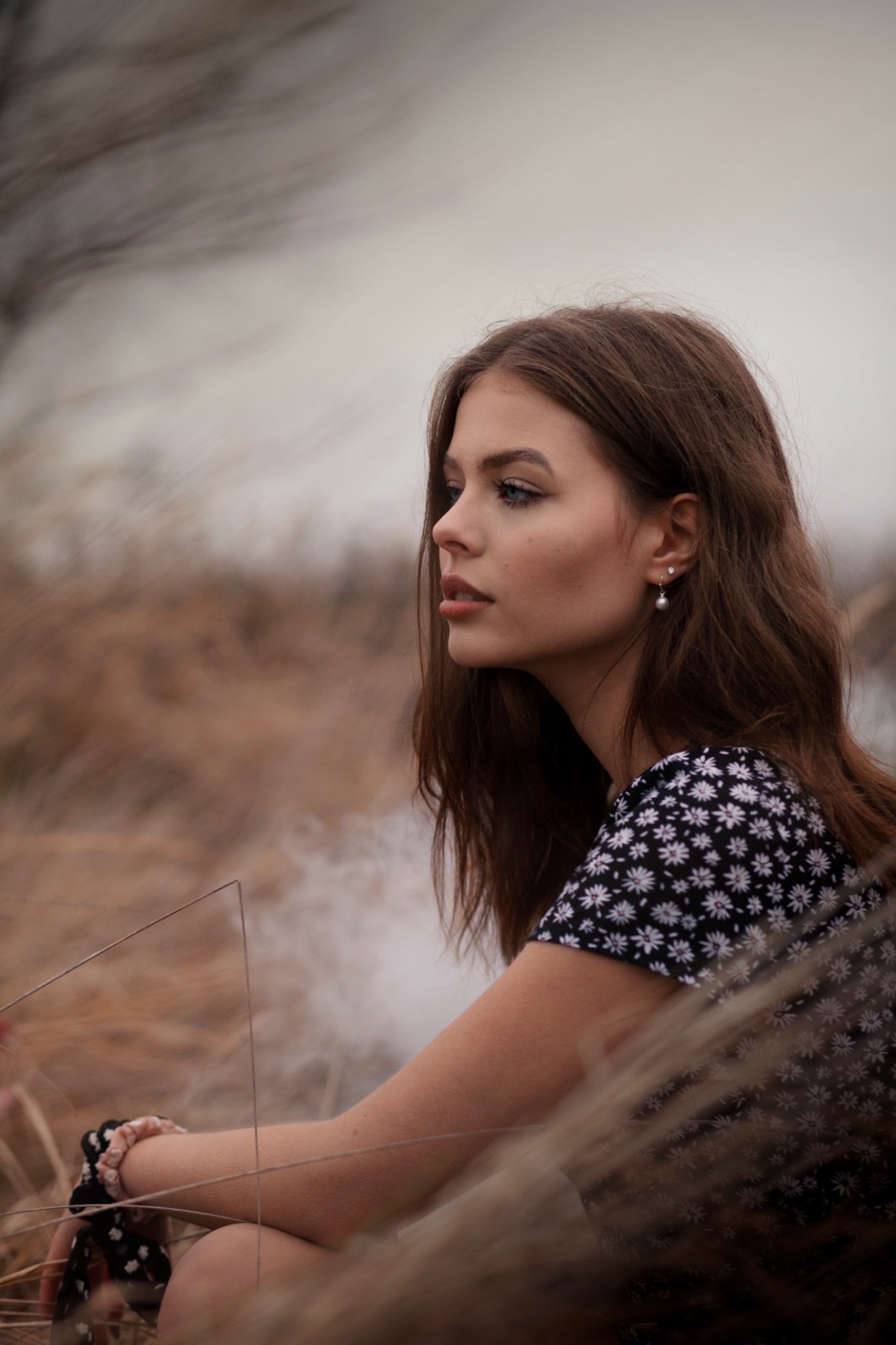Portret młodej kobiety z profilu, w sukience w kwiaty, z kolczykami z perłami, siedzącej w trawie z dymem w tle.
