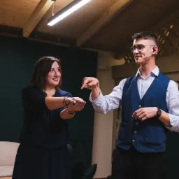 Młody mężczyzna w okularach i niebieskiej kamizelce, z mikrofonem nagłownym, demonstruje trik z kartami kobiecie w granatowej sukience podczas występu na tle zielonej ściany.