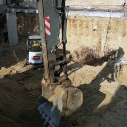 Koparka Bobcat z łyżką w wykopie fundamentowym, widoczne zbrojenia i ściany wykopu z elementami konstrukcyjnymi budynku w tle.