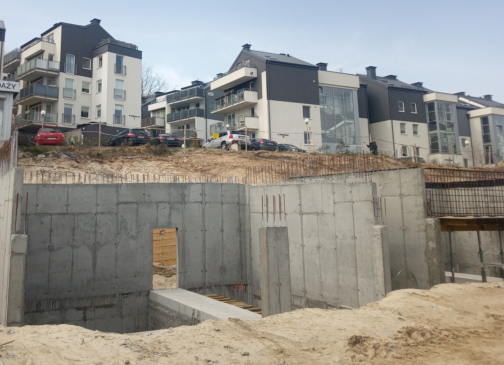 Wykop pod fundamenty budynku wielorodzinnego z widocznymi betonowymi ścianami szalunkowymi i wystającymi prętami zbrojeniowymi, na tle nowej zabudowy mieszkalnej.