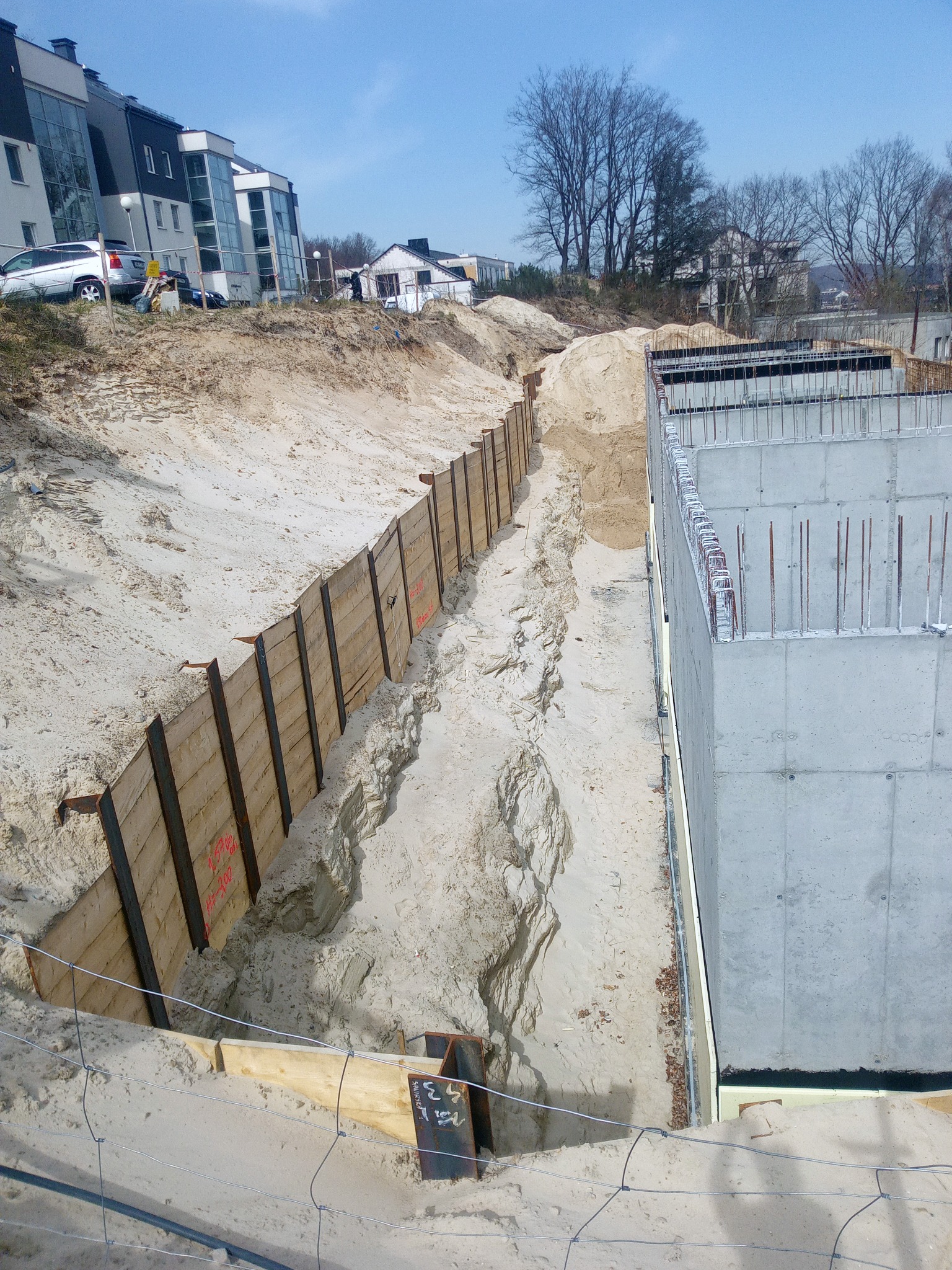 Wykop zabezpieczony deskowaniem pionowym z metalowymi profilami, obok betonowa konstrukcja z wystającymi prętami zbrojeniowymi, piaszczyste podłoże, w tle zabudowa mieszkalna.