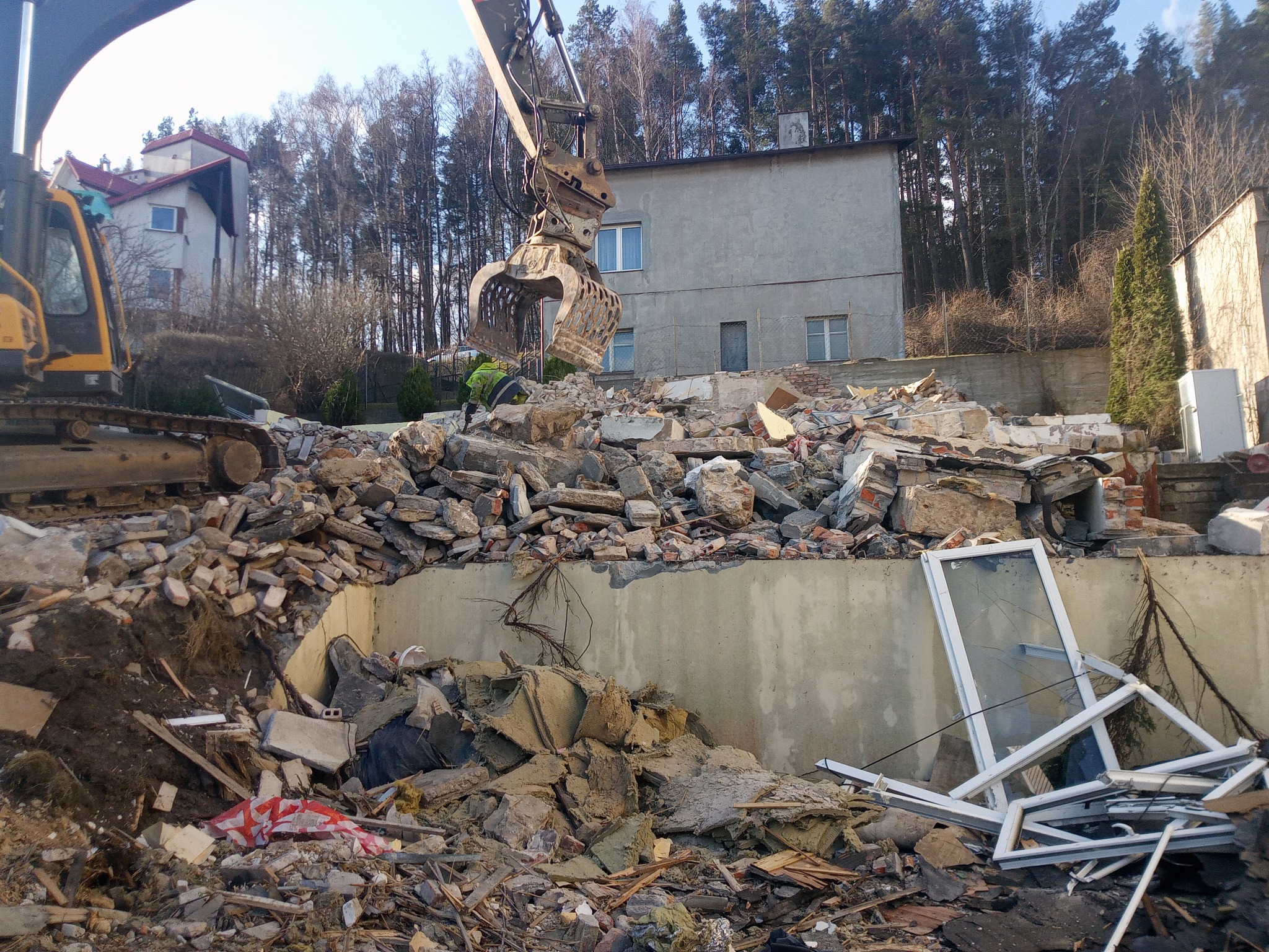Żółta koparka z chwytakiem rozbiera gruzowisko po zburzonym budynku, w tle budynek mieszkalny i drzewa.