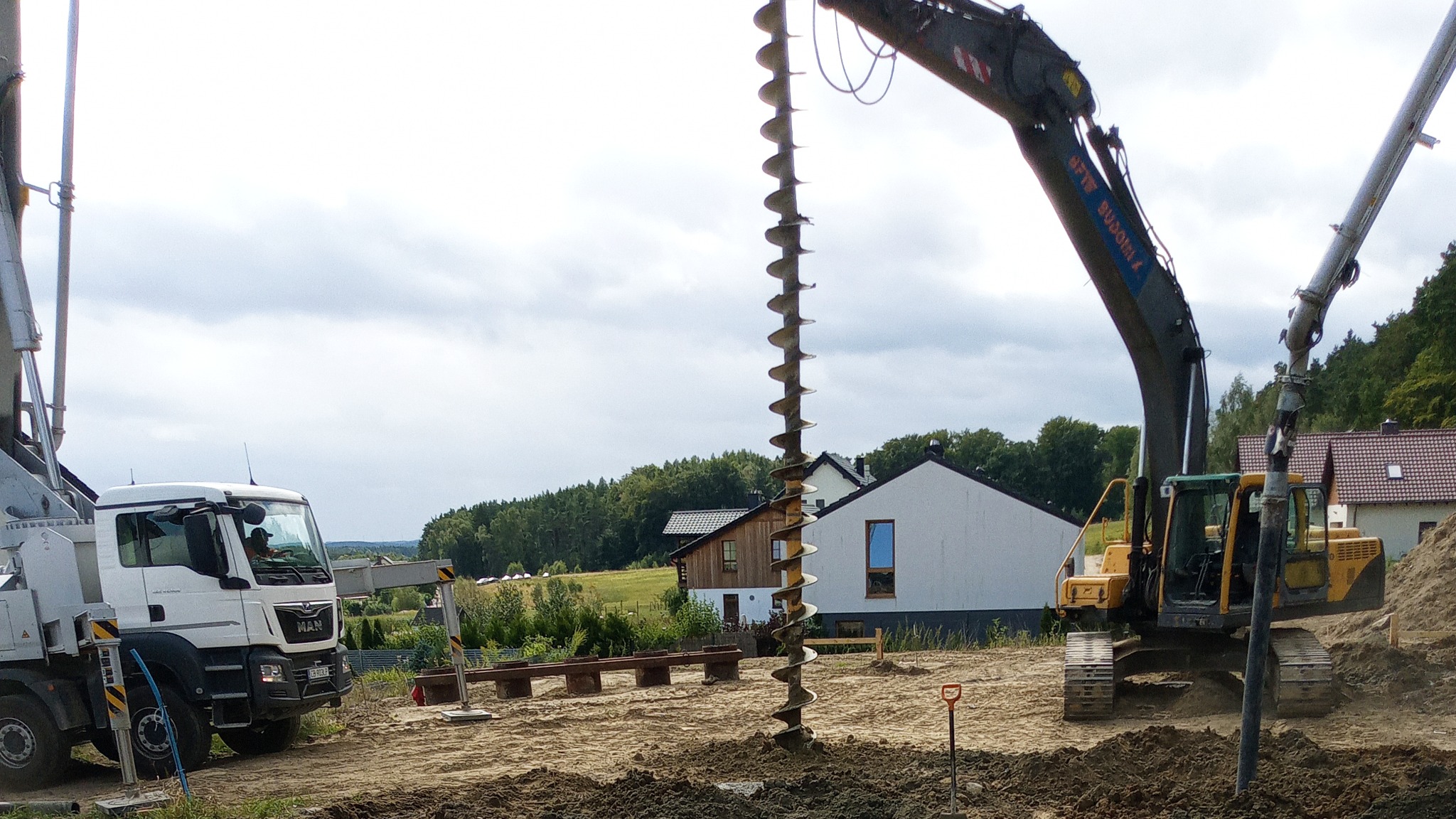 Żółta koparka z wiertłem ślimakowym w trakcie pracy na piaszczystym terenie budowy, w tle biały budynek mieszkalny i las, obok stoi biała ciężarówka MAN.