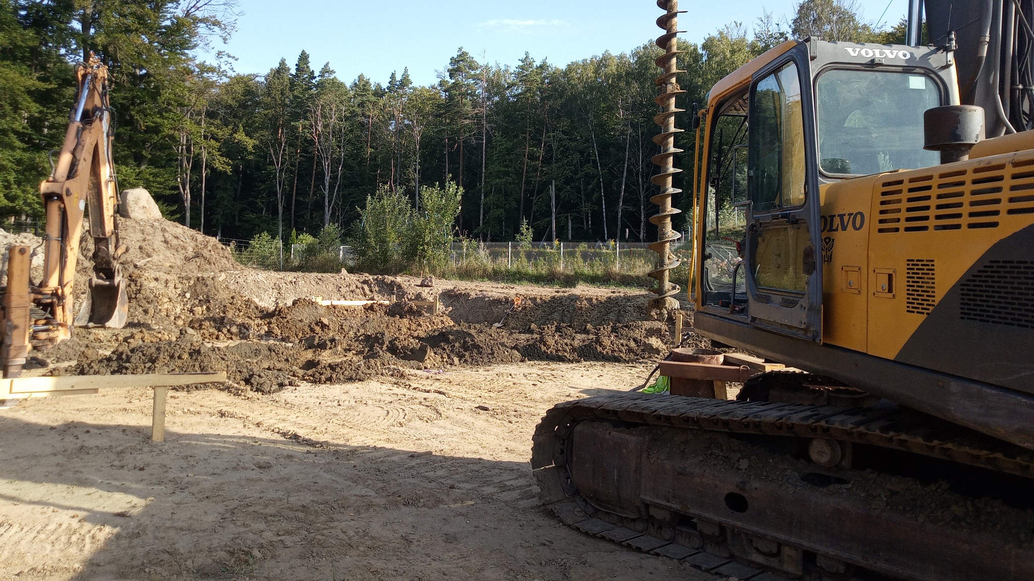 Żółta wiertnica Volvo z wiertłem ślimakowym na placu budowy, widoczny wykop i stos ziemi w tle las