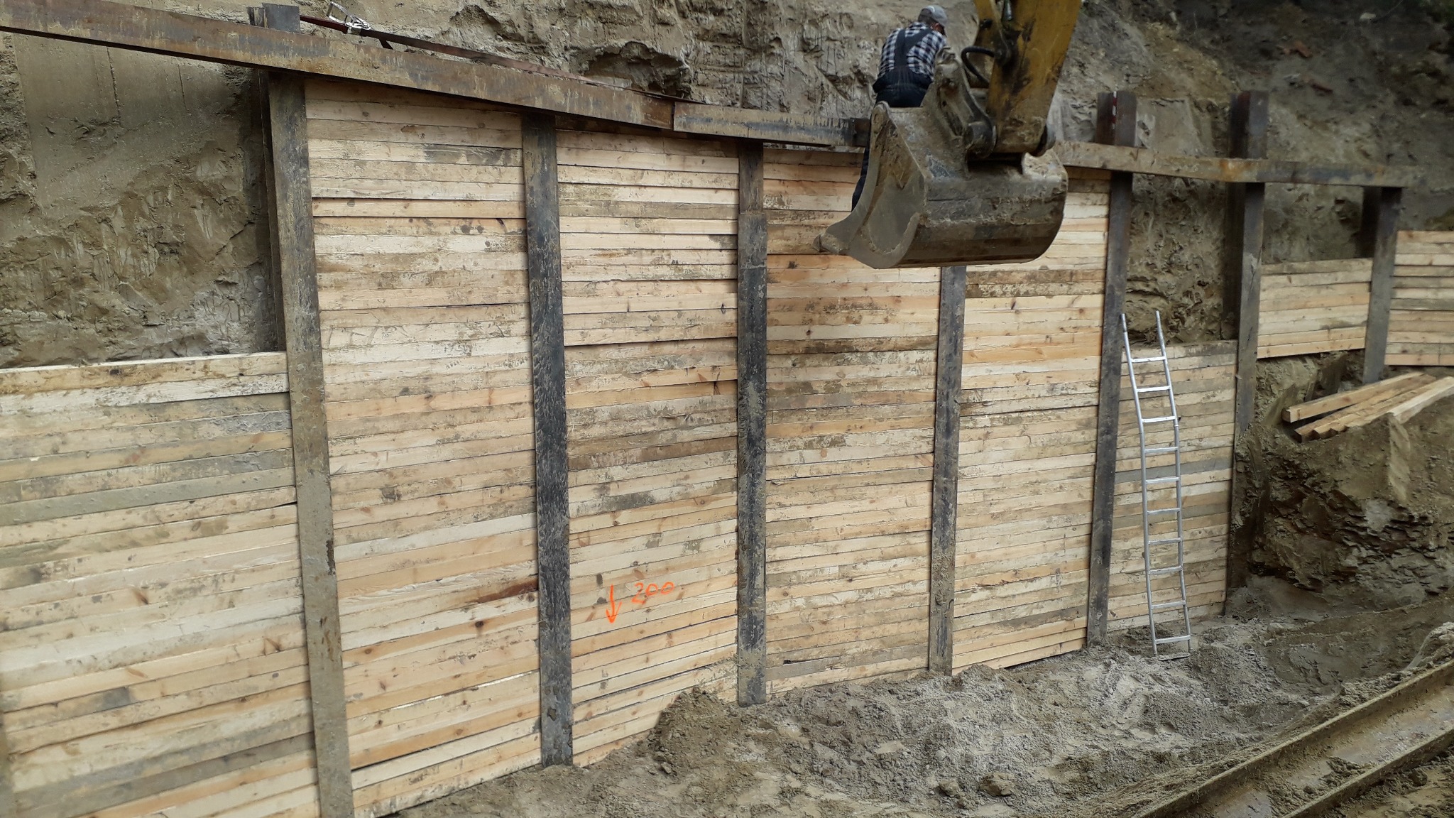 Ściana wykopu zabezpieczona drewnianą konstrukcją z metalowymi słupkami, widoczna koparka z operatorem w górnej części kadru i drabina oparta o ścianę wykopu.