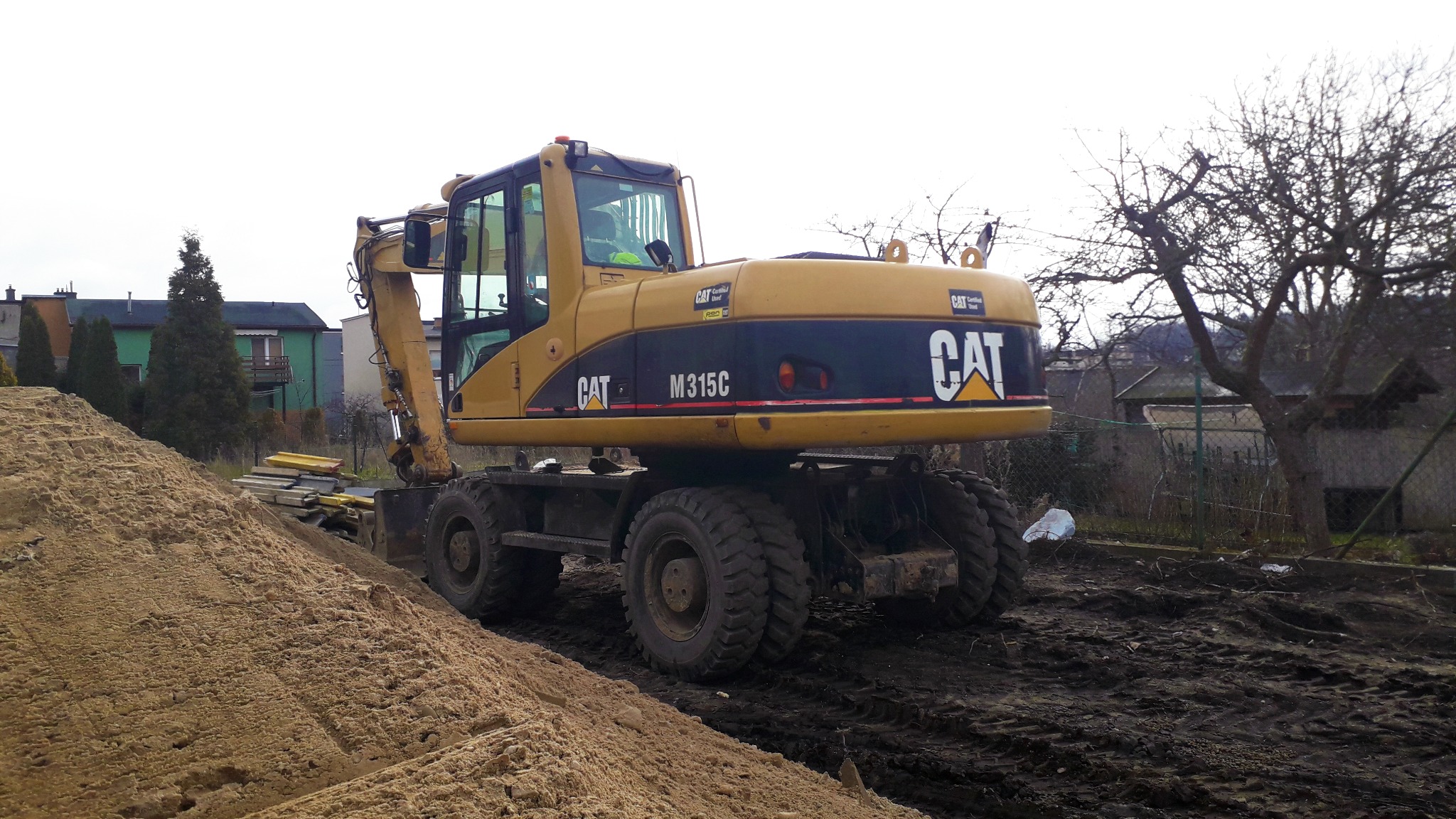 Żółta koparka kołowa CAT M315C na błotnistym terenie budowy, obok sterta piasku i ułożone płyty chodnikowe, w tle zabudowa jednorodzinna.
