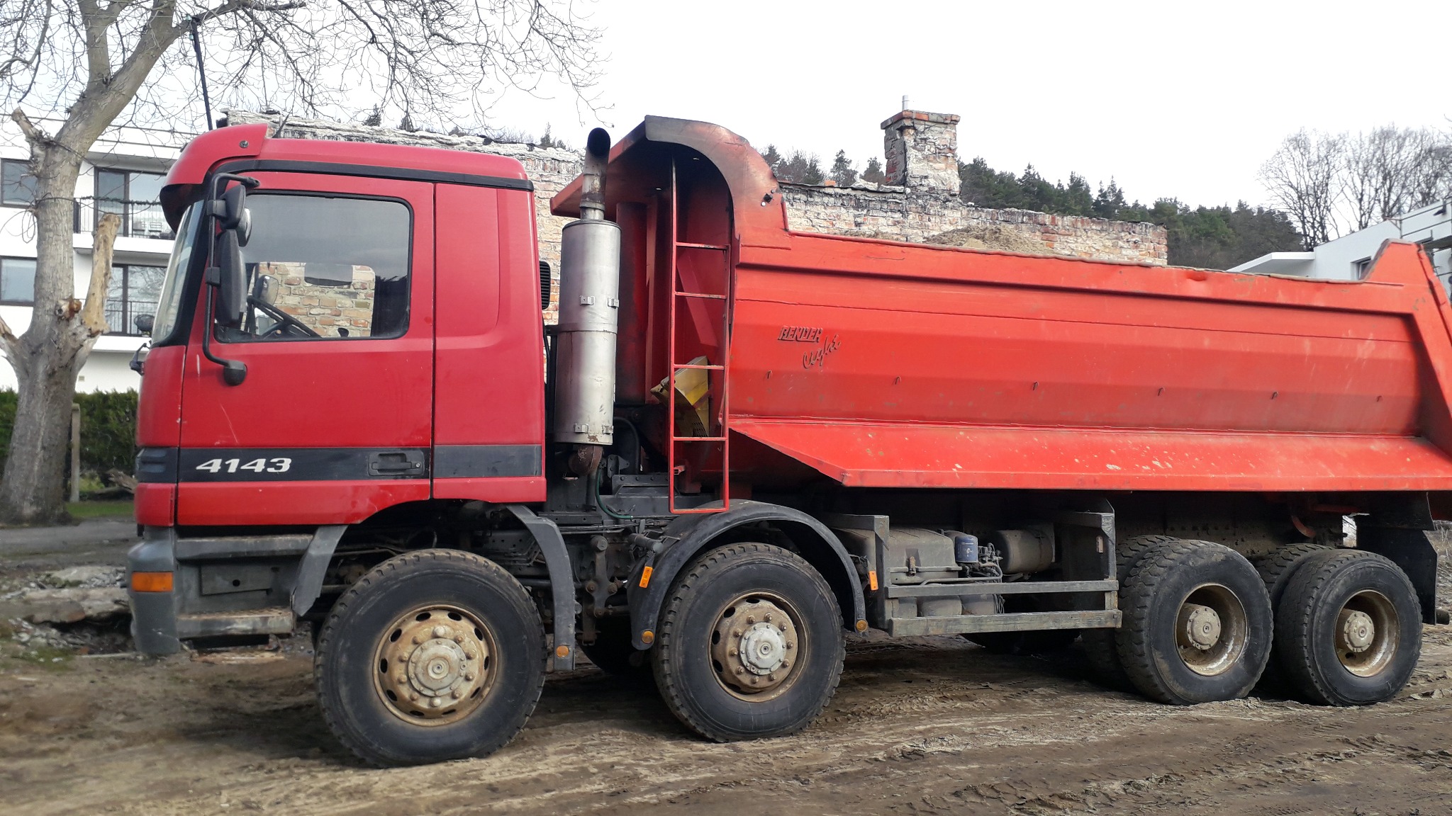 Czerwona wywrotka MAN 4143 na błotnistym terenie, z widocznym budynkiem w tle i fragmentami gruzu.