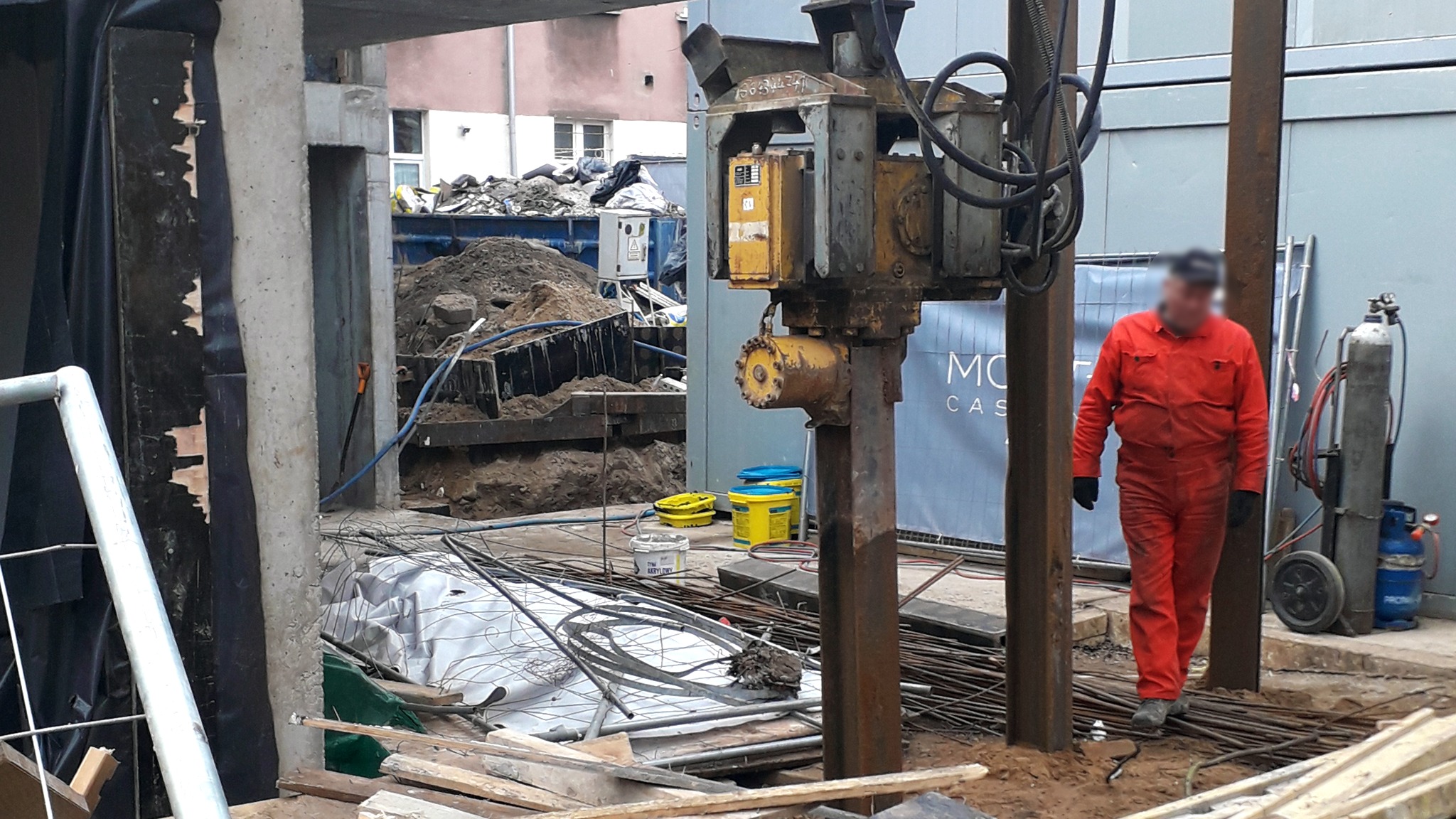 Pracownik w czerwonym kombinezonie obsługuje maszynę do wbijania pali w ziemię na placu budowy, widoczne wykopaliska, stalowe pręty i butle z gazem.
