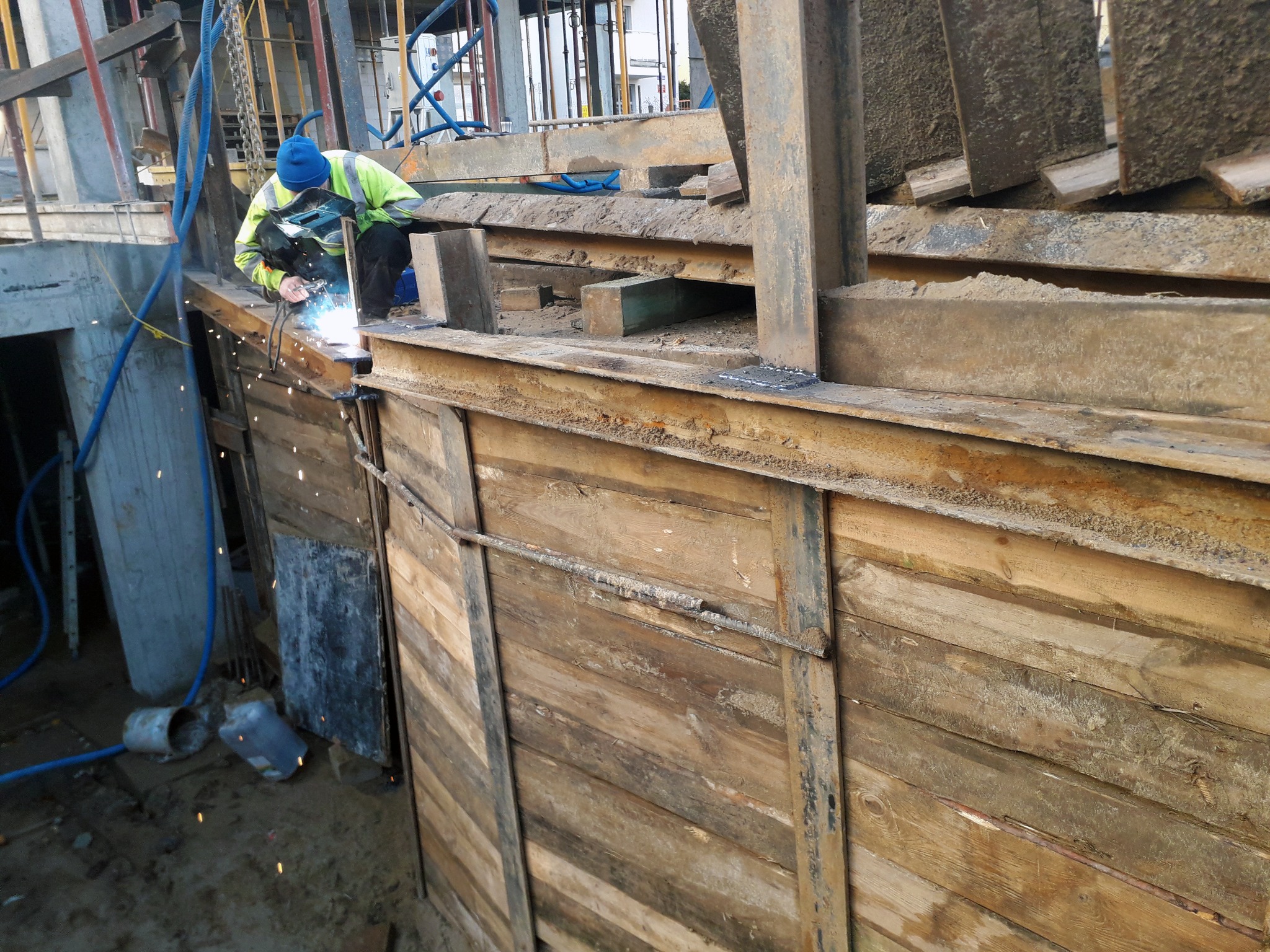 Spawacz w niebieskiej czapce i żółtej kamizelce spawa metalową konstrukcję szalunku wykopu, iskry rozświetlają drewniane deski i metalowe elementy, widoczne węże i elementy rusztowania w tle.