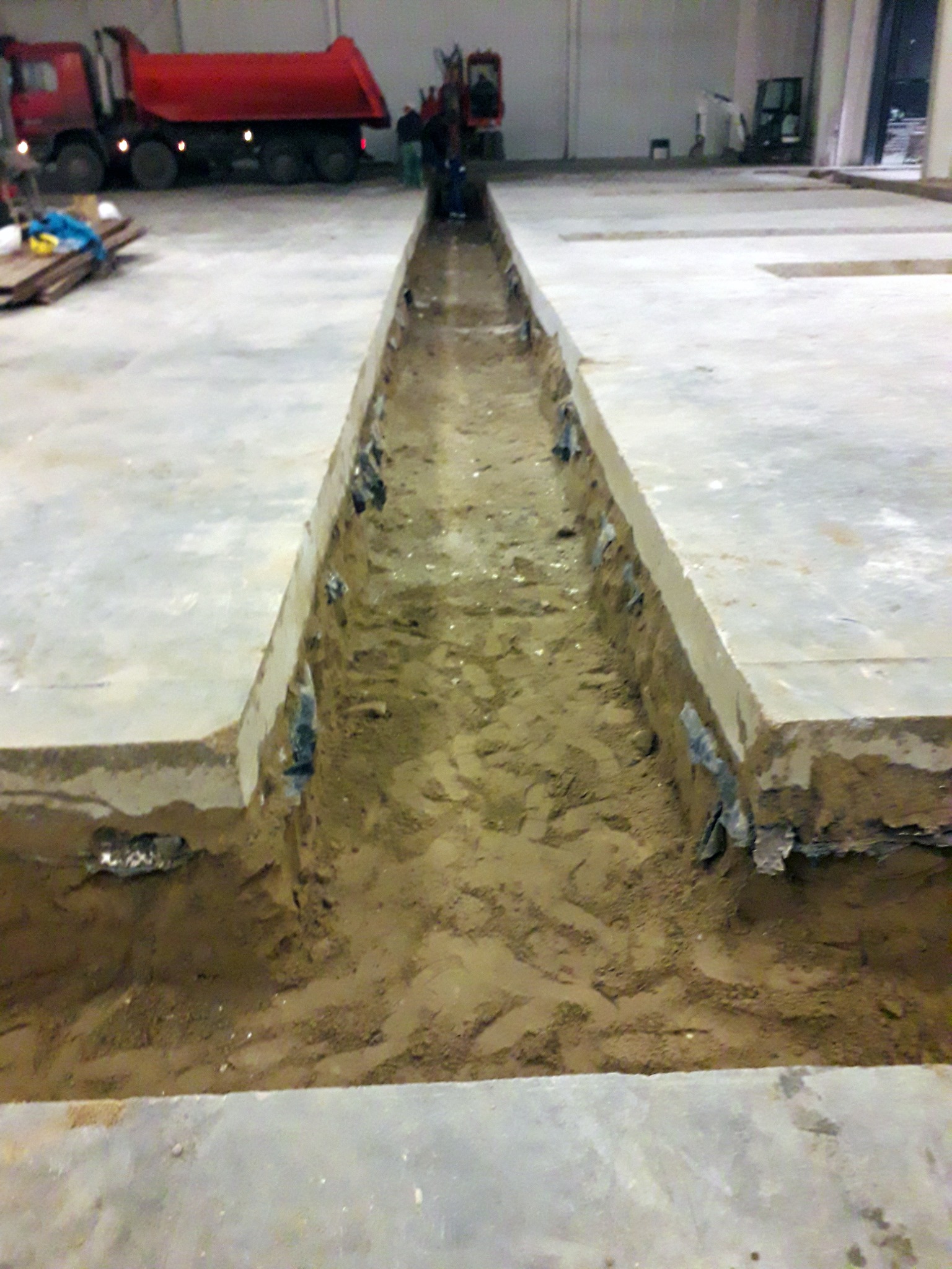 Wykop w kształcie litery V wewnątrz budynku z betonową posadzką, widoczny czerwony samochód ciężarowy w tle.