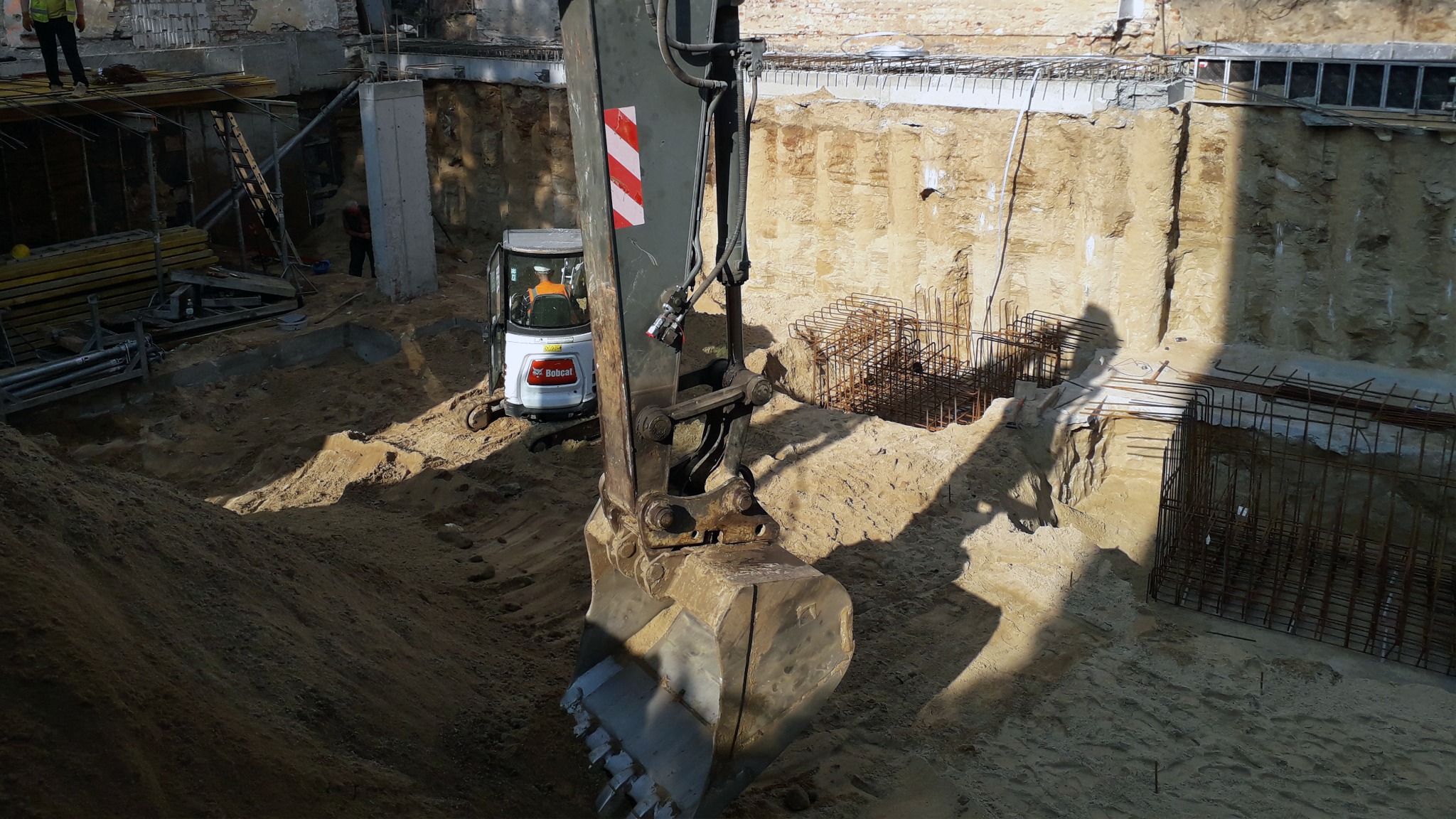 Koparka Bobcat z łyżką w wykopie fundamentowym, widoczne zbrojenia i ściany wykopu z elementami konstrukcyjnymi budynku w tle.