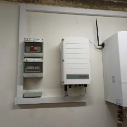 Biała skrzynka z bezpiecznikami, falownik z anteną i piec grzewczy zamontowane na ścianie w pomieszczeniu technicznym. Widoczne rurki instalacyjne.