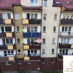Fasada bloku mieszkalnego z panelami słonecznymi na balkonie, widok z góry, z logo firmy w prawym dolnym rogu.