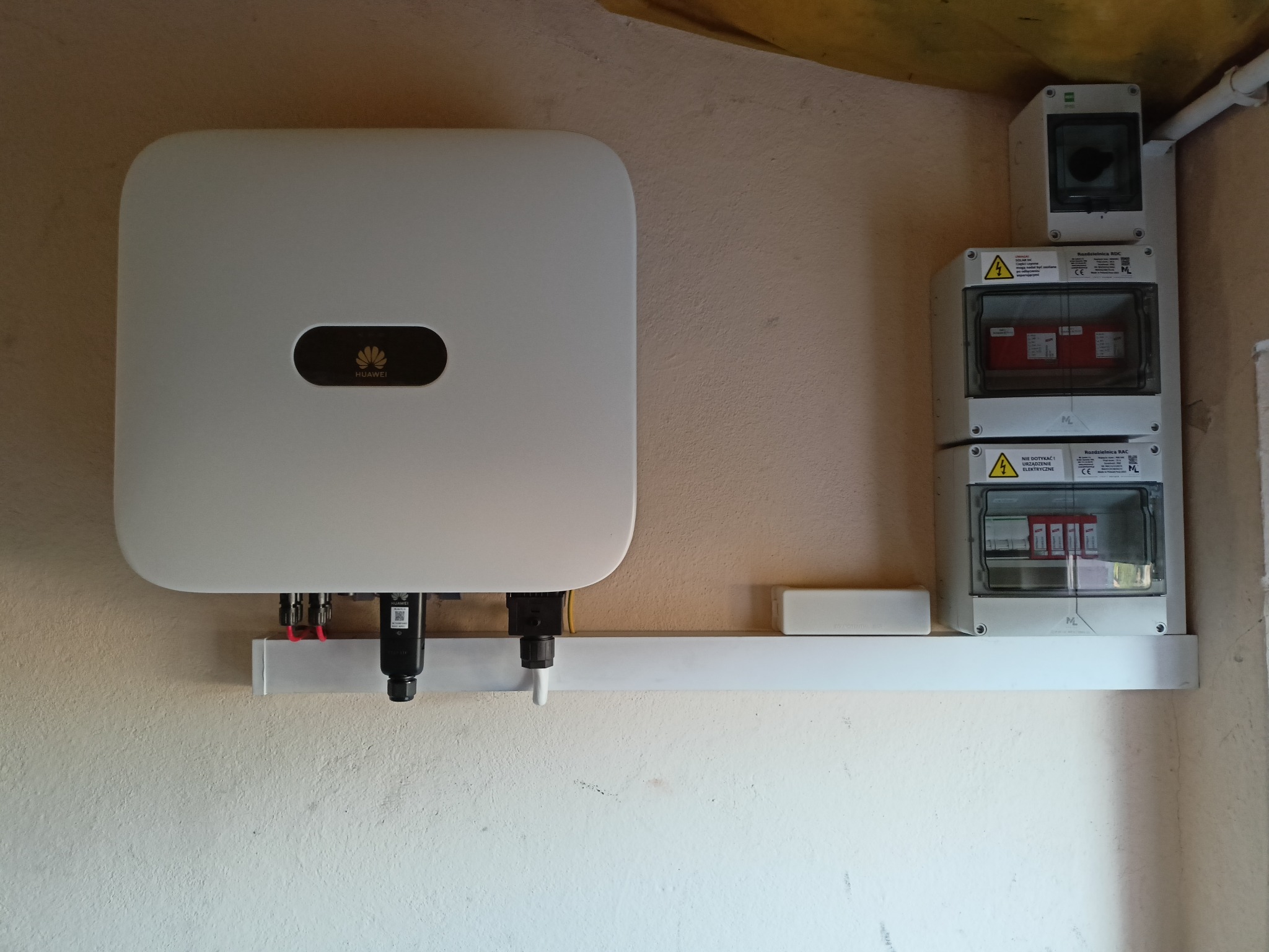 Biały falownik marki Huawei zamontowany na ścianie, obok skrzynka z bezpiecznikami i wyłącznik prądu. Instalacja wewnątrz budynku.