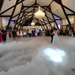 Para Młoda tańczy pierwszy taniec w chmurach na tle gości weselnych i dekoracyjnego sufitu z drewnianą konstrukcją oraz kryształowymi żyrandolami.