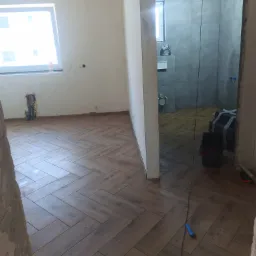 Układanie drewnopodobnych płytek w jodełkę w pokoju i łazience w trakcie remontu, widoczne narzędzia i instalacje.